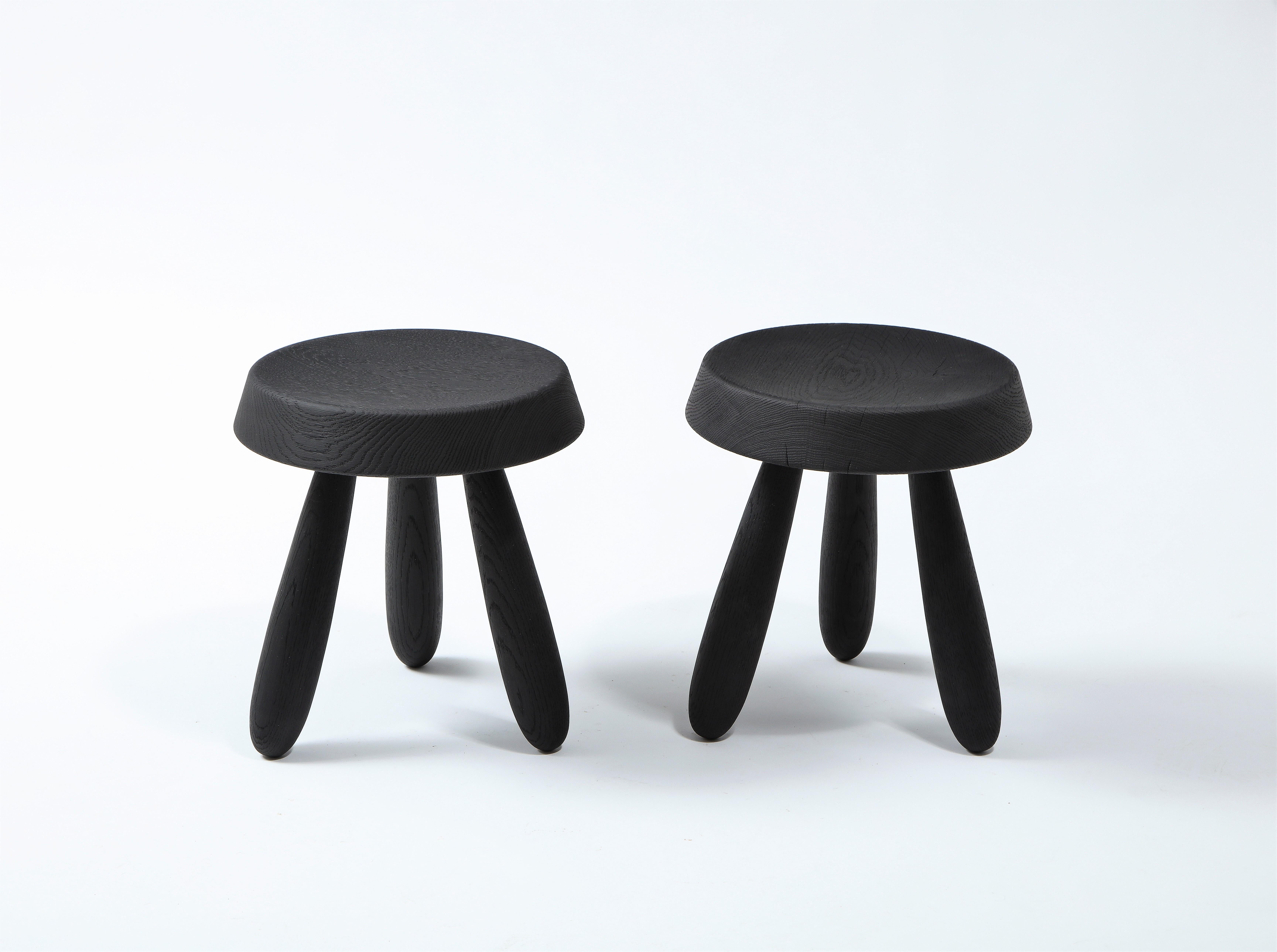 A pair of beautifully crafted tripod stools by French designer Douglas Mont and produced by Facto Atelier Paris. A thick beveled seat sits on three spindle shaped feet. Burnt Oak finish with visible wood grain. Appearance, cracks and grain may