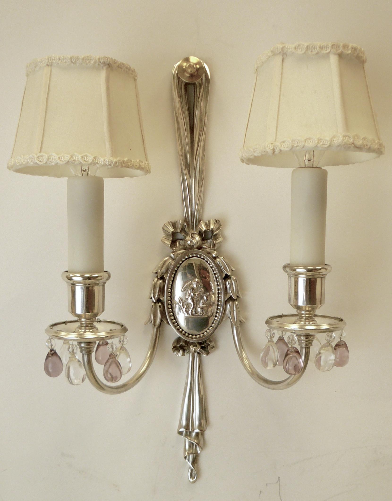 This handsome pair of sconces feature oval repousse Classical style reserves, and drapery, bow, and bellflower motifs. The twin arms are hung with clear and pale amethyst crystal drops.
