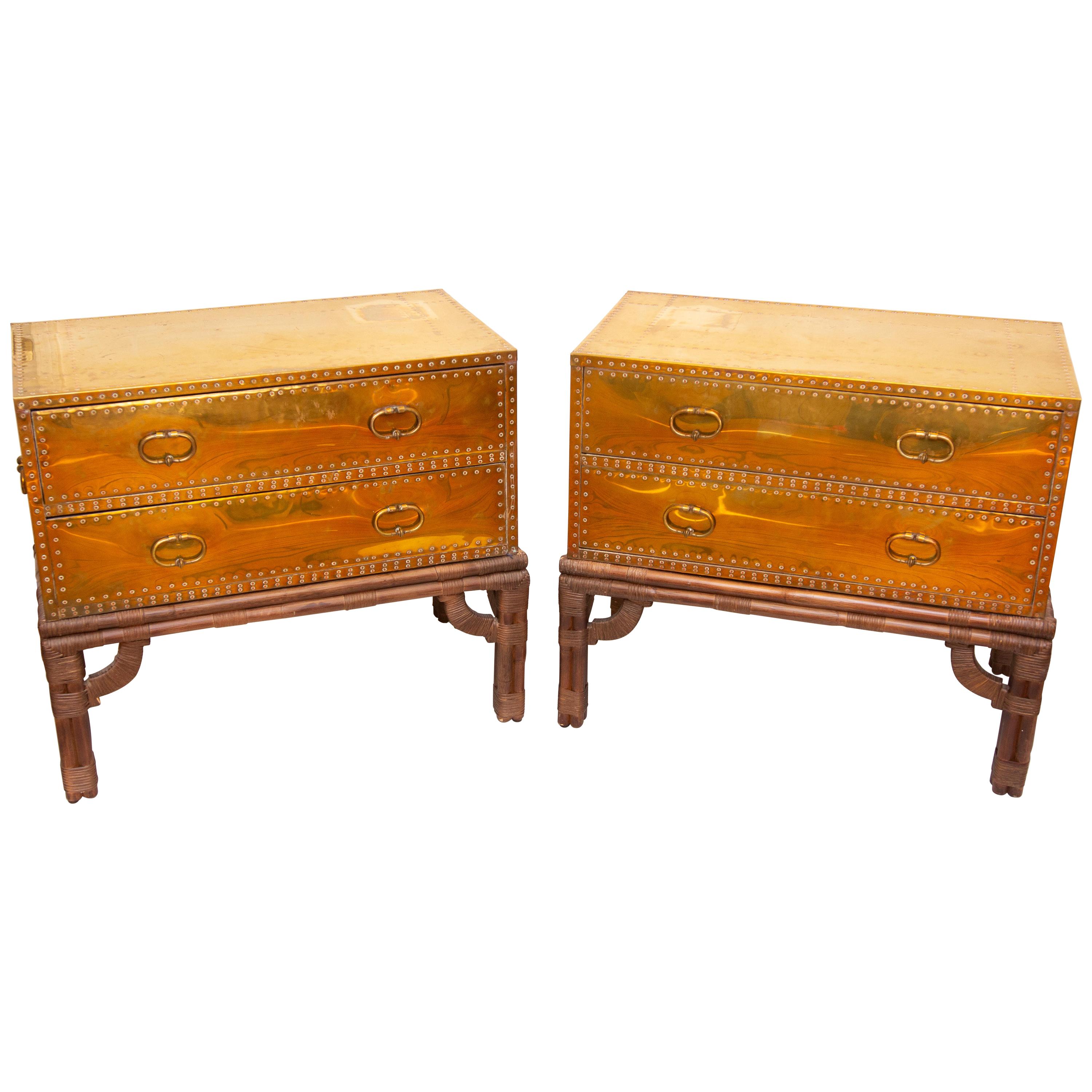 Pair of Campaign style two-drawer chests on the original rattan stands. Heavy riveted brass over wood. Very well made and sturdy. Backs are also clad. They would work well as commodes, consoles or end tables, circa 1970s by Sarreid Ltd. They also