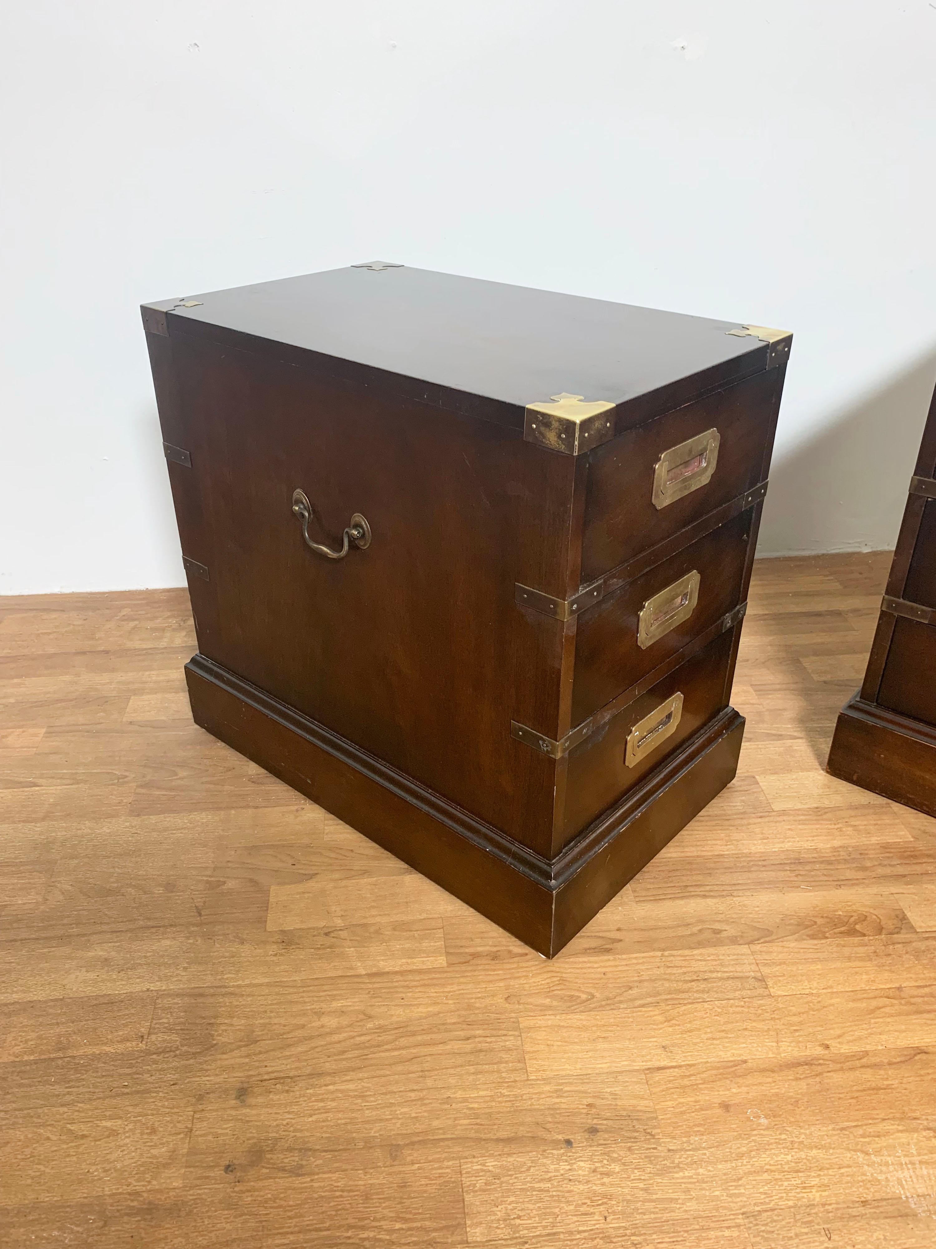 A pair of British made three drawer campaign style chests in mahogany, featuring brass corners, strapping on all three drawers, baseboard molding and working lift handles. Finished backs make these a great side / end table option; also perfectly