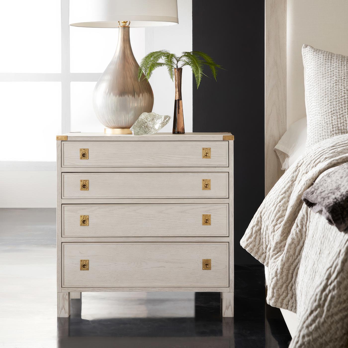 Drawing inspiration from classic campaign furniture, this refined bedside chest combines traditional military aesthetics with contemporary craftsmanship. The white-painted oak construction features a carefully wire-brushed finish that emphasizes the