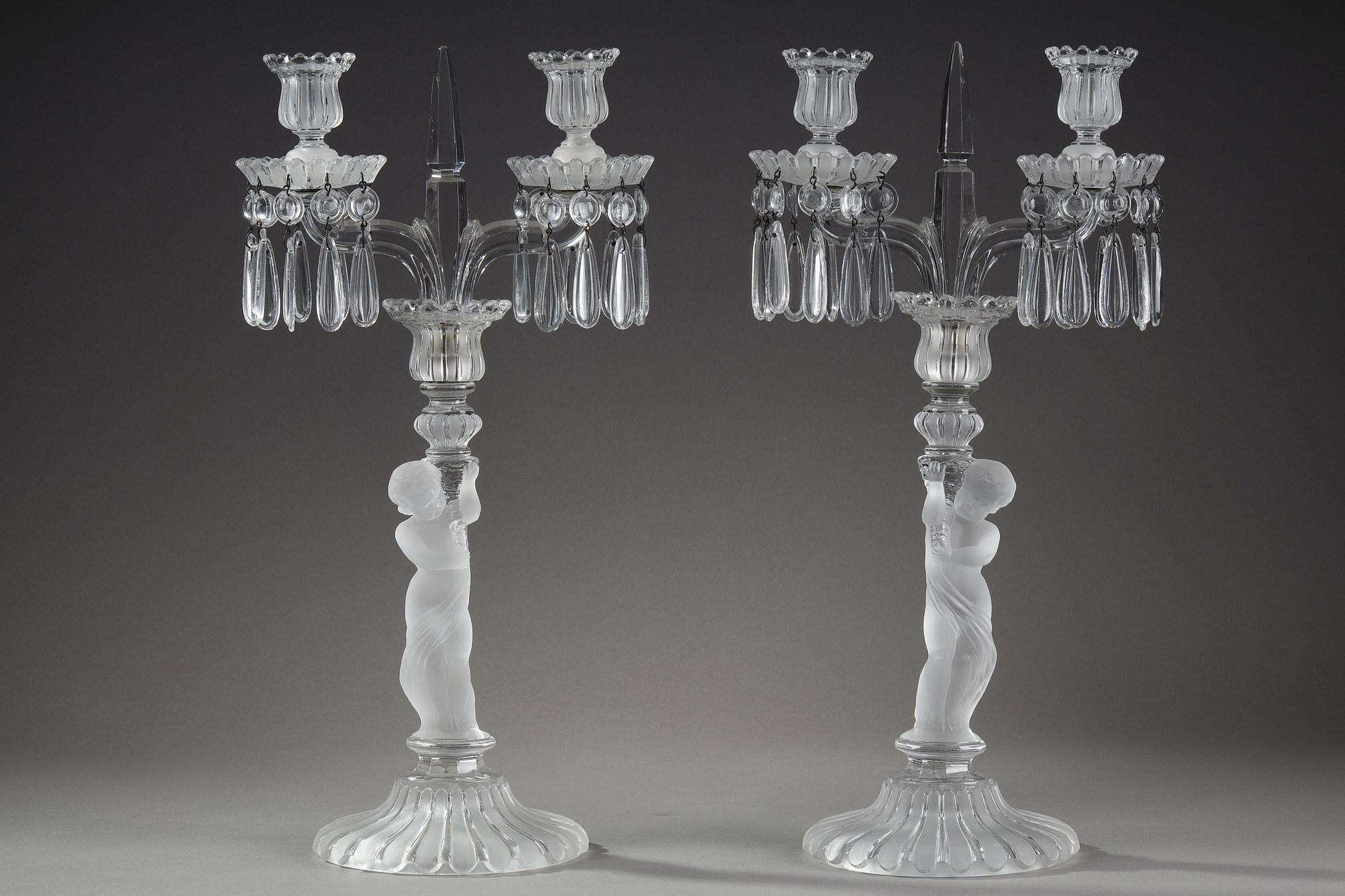 Paire de candélabres en cristal clair et satiné de Baccarat avec deux bras de lumières ornés de pampilles. Au centre s'élève un motif de poignard. La tige représente un putto tenant une corne d'abondance. La base et les bobèches sont décorées de