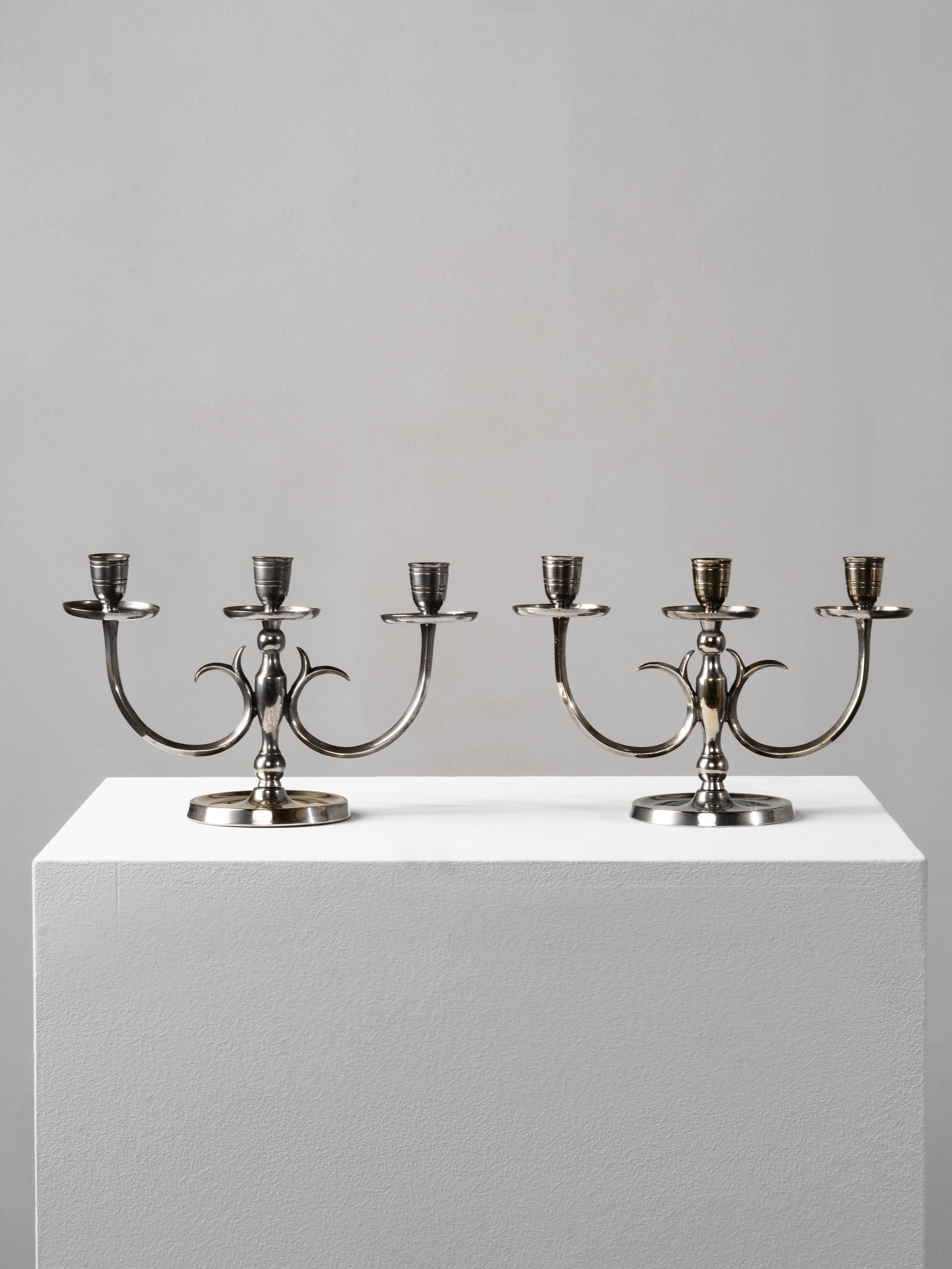 Una raffinata coppia di candelabri Model 8013 disegnati da Paavo Tynell e prodotti da Taito Oy negli anni Trenta. Realizzati in ottone nichelato, questi eleganti portacandele a tre bracci rappresentano la prima espressione del linguaggio del design