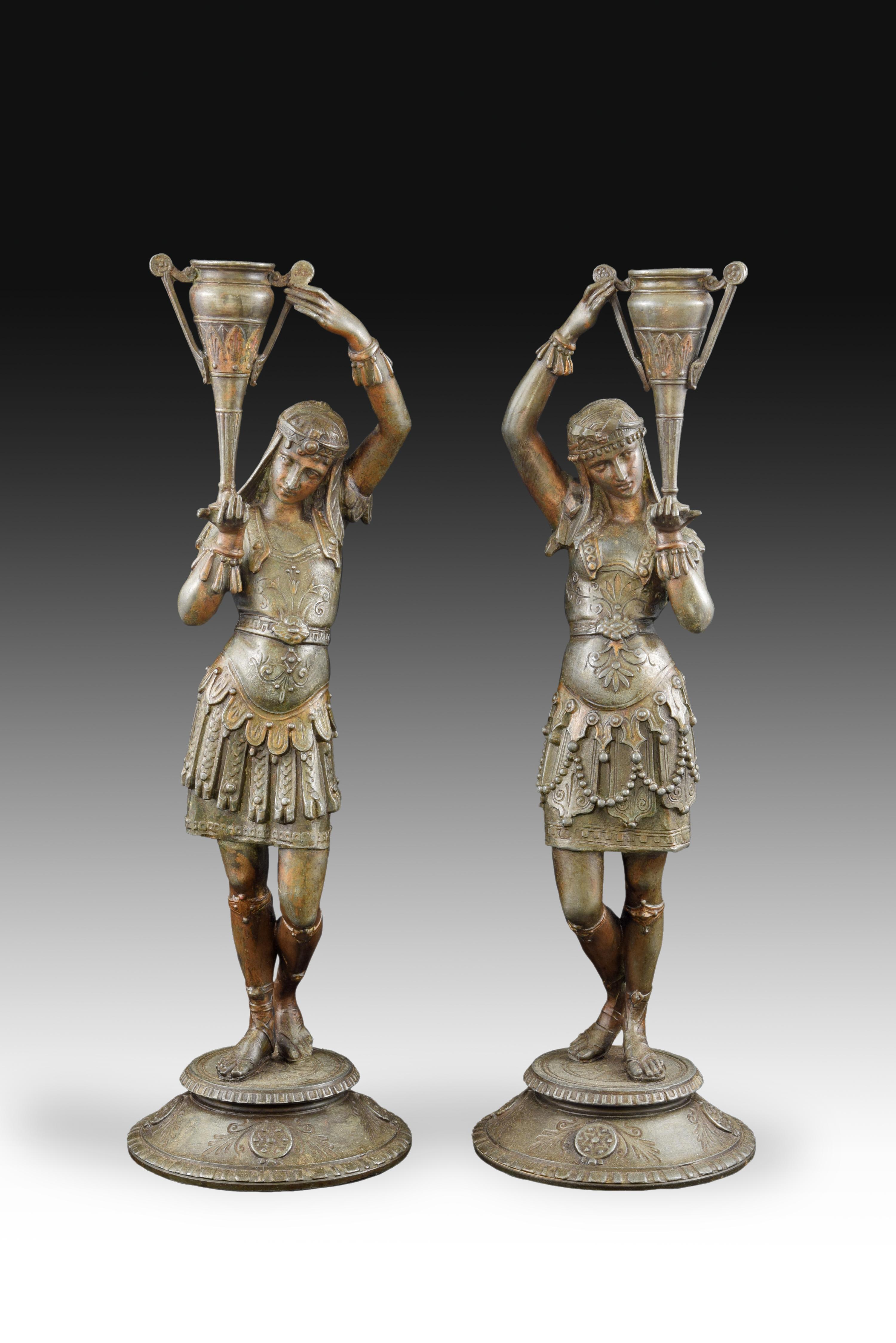 Pareja de candeleros realizados en calamina que conservan restos de policromía y tienen bases circulares sobre las que se sitúan dos figuras vestidas a la romana según la visión romántica del siglo XIX, cada una de las cuales sostiene un jarrón