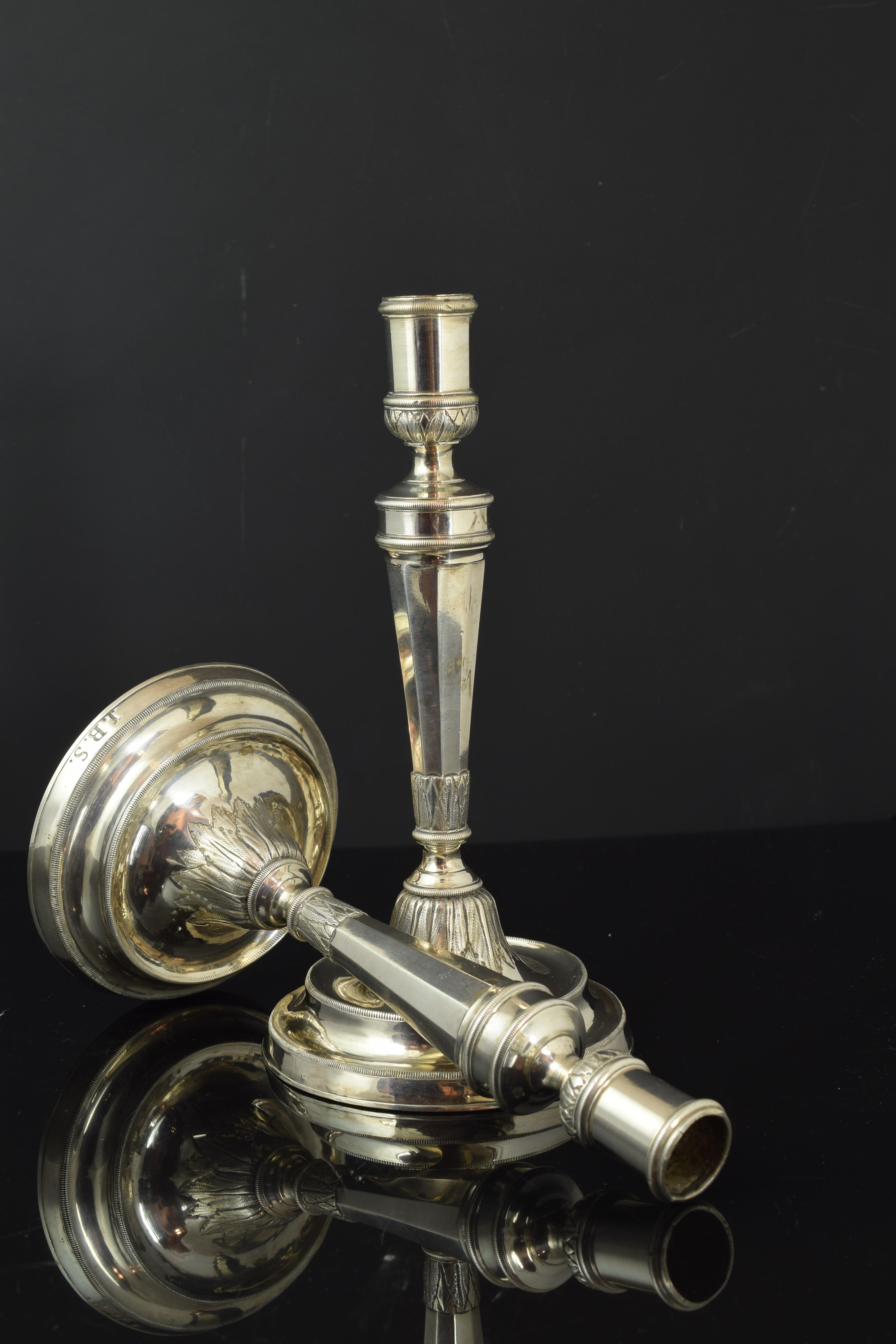 Pair of candlesticks with a circular base, in which initials are presented, with a column-shaped axis with a bell-shaped base decorated with leaves in slight relief. The shapes and moldings that decorate them respond to the influence of