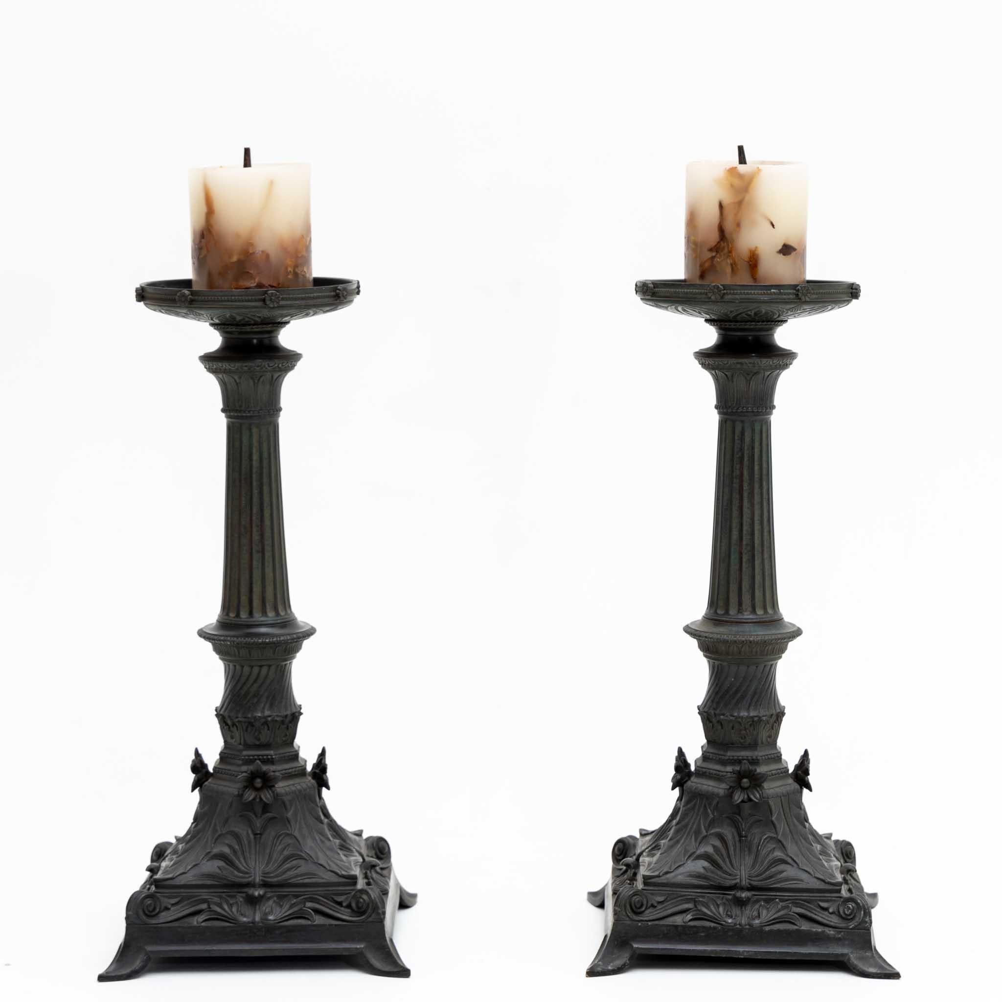 Pareja de candeleros de metal con pie cuadrado con adornos de hojas estilizadas y decoración floral aplicada alrededor del fuste y la bandeja recogegotas. El eje también está estriado y perfilado.