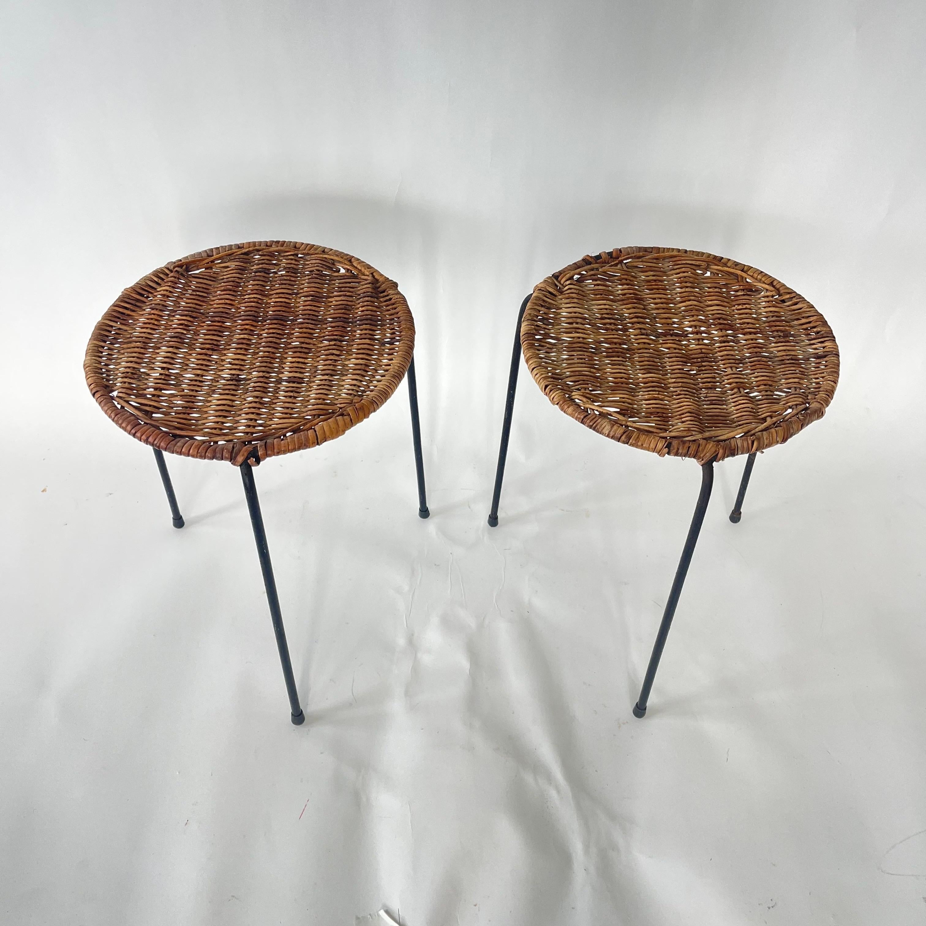 Coppia di tavoli o sgabelli impilabili a treppiede attribuiti a Carl Auböck.  Realizzate con telai in ferro nero forgiato con tre gambe che sostengono un piano o una seduta in rattan intrecciato.  Attribuito a Carl Auböck, Austria, anni '50 circa.