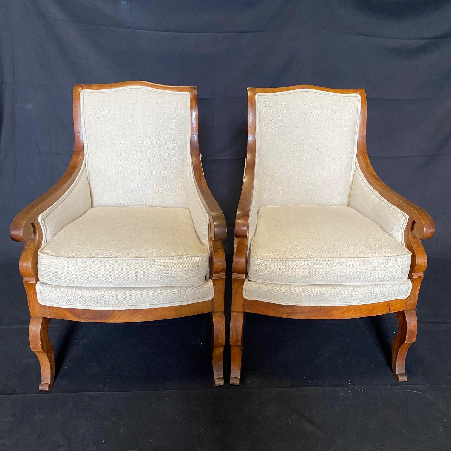 Pair of antique French Empire walnut armchairs or bergères havingtailored framework accented by a subtly arched seat back and curved arm rests that terminate in a graceful joinery just above the beautifully carved legs. New upholstery in high