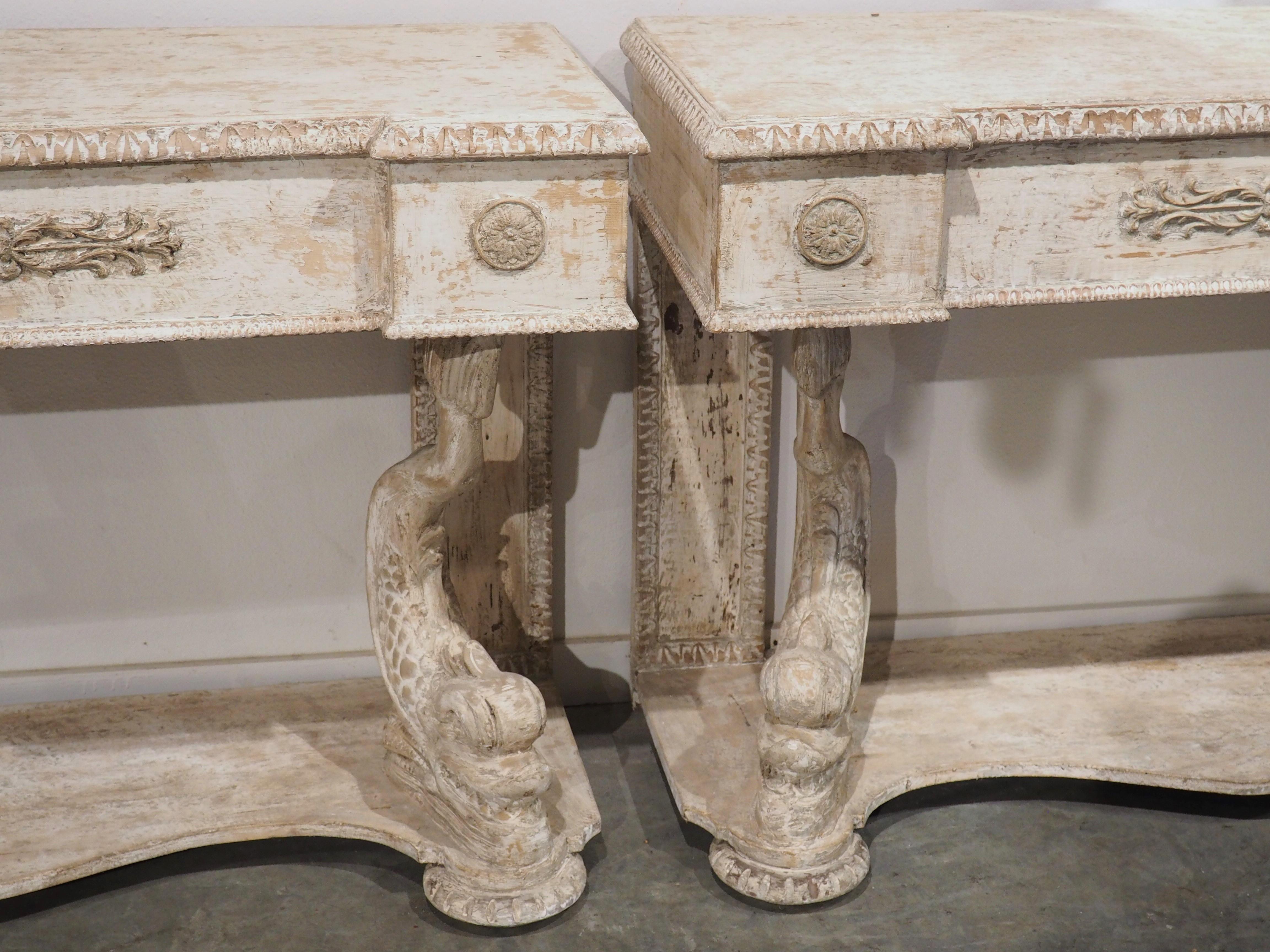 hand carved in Florence, Italy, and masterfully painted and distressed, this pair of console tables feature fantastically detailed dolphin supports. Both consoles have been parcel painted cream with beige and gray areas, while the angular tops have