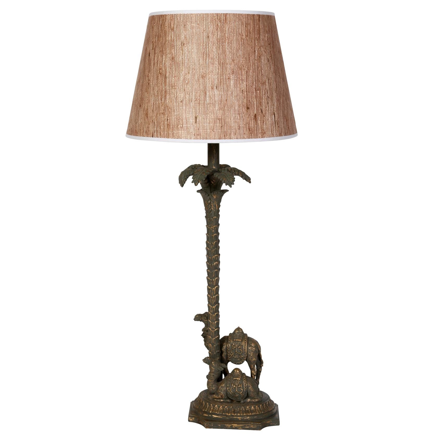 A pair of carved and painted table lamps in the form of camels at the base of a palm tree.  The intricately carved lamps feature one standing camel and one sitting camel with a tall palm tree between them.   A detailed saddle sits on the back of