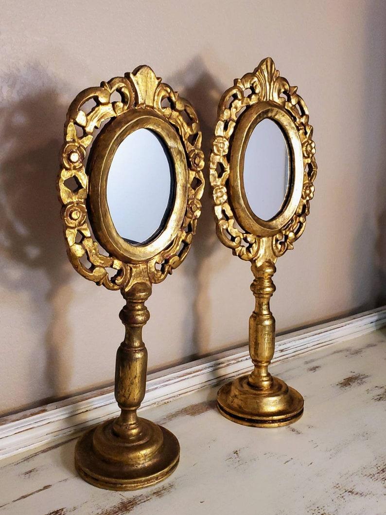 A striking pair of antique hand carved gilded solid wood table mirrors. Inspired by Italian design, this magnificent Mexican folk art features elegant oval shaped mirrors with brilliant gold gilt (dore bronze) finish, intricate piecered scrolling