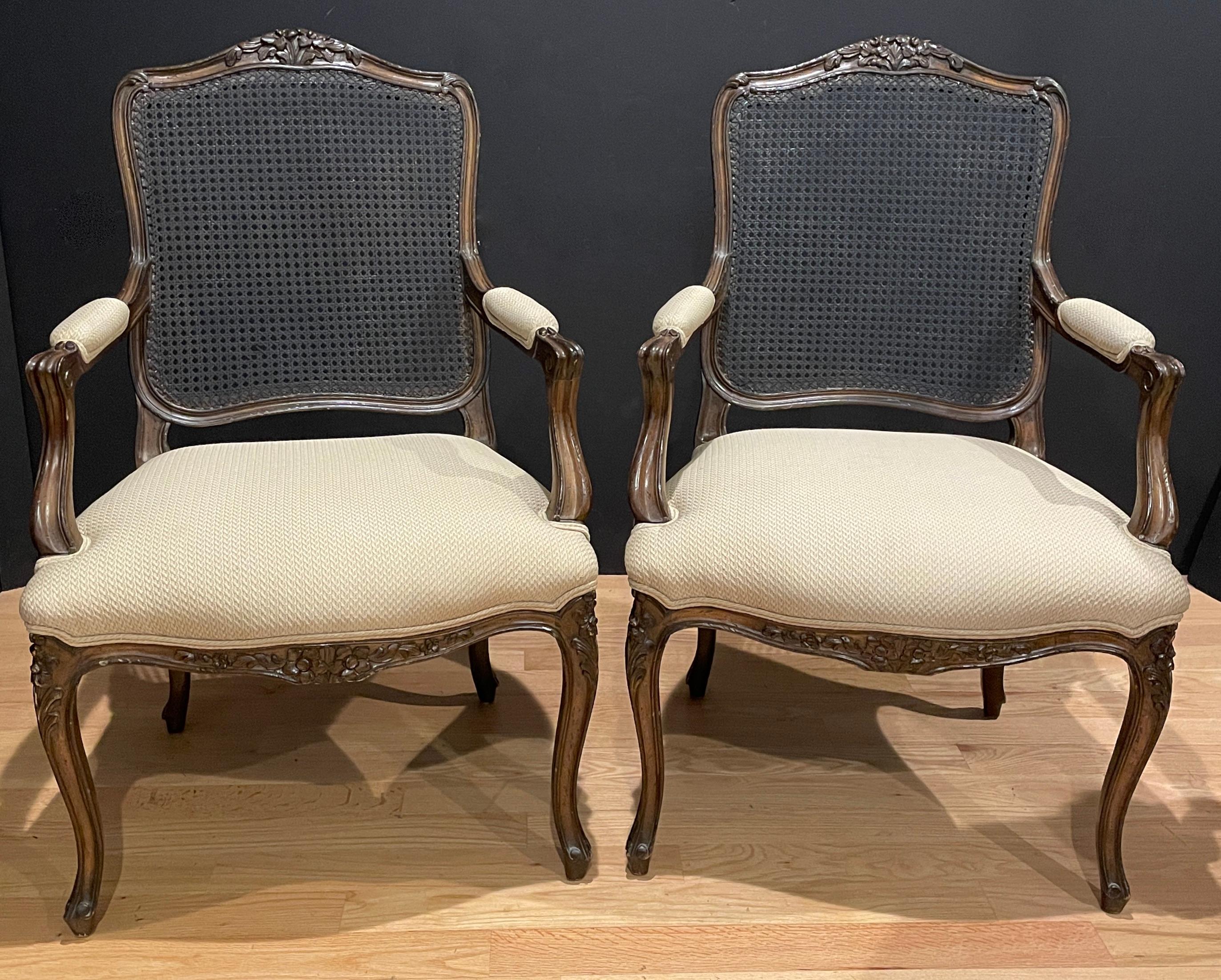 Pair of carved, caned and upholstered Louis XV style armchairs with light tan upholstery. Caned back and upholstered seat and arm pads. Floral carvings with walnut finished beechwood.