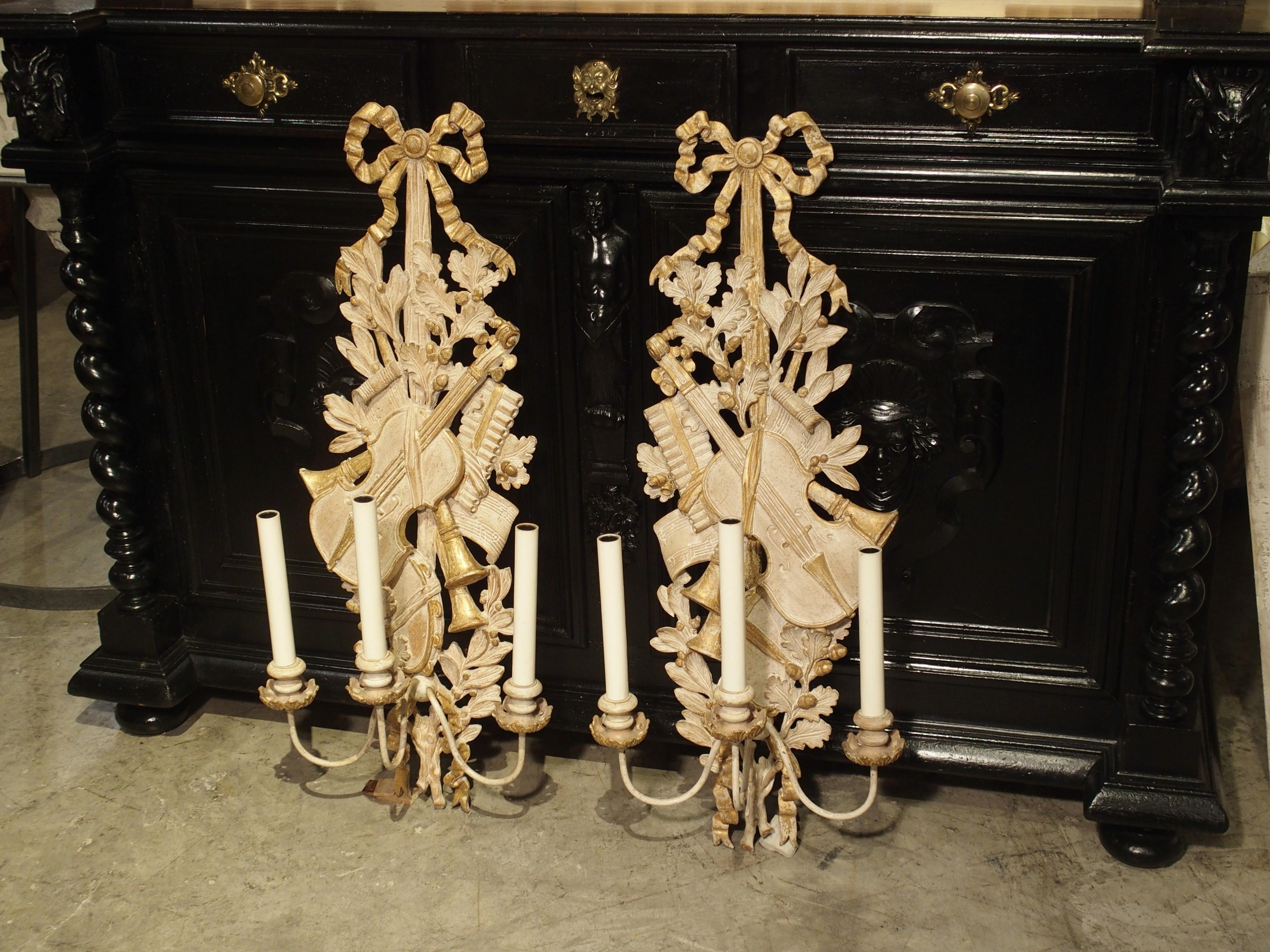 This pair of French sconces has been carved out of Linden wood, in the Louis XVI style. The 3-armed sconces are painted in a light cream and gold, and the carvings represent musical instruments, laurel branches, and oak leaves with acorns, all hung