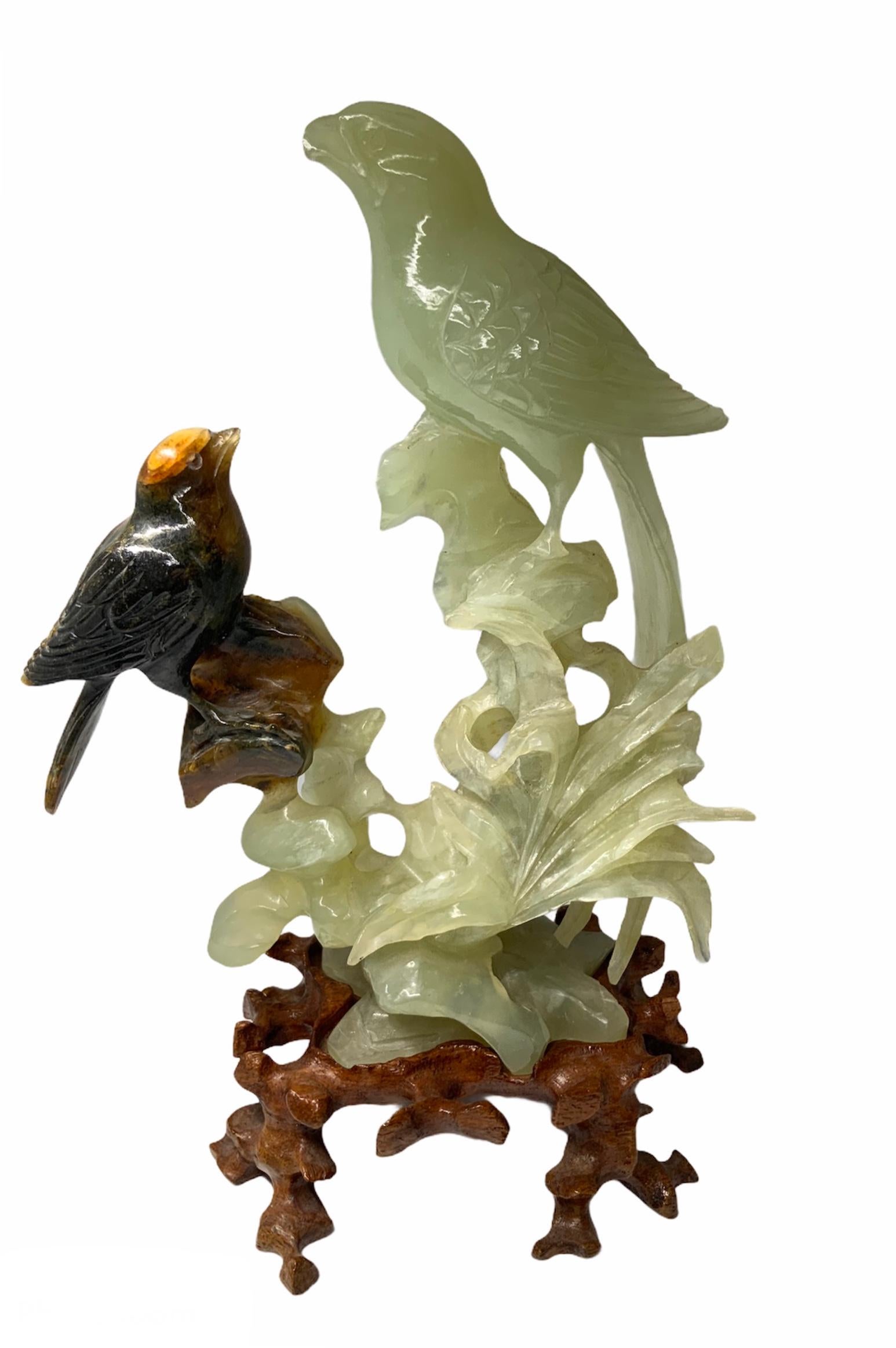 These are a pair of carved russet jade bird and jade parrot sculptures. They are standing up in some branches. The whole jade sculpture is standing over a carved wood base that appears like cut branches.