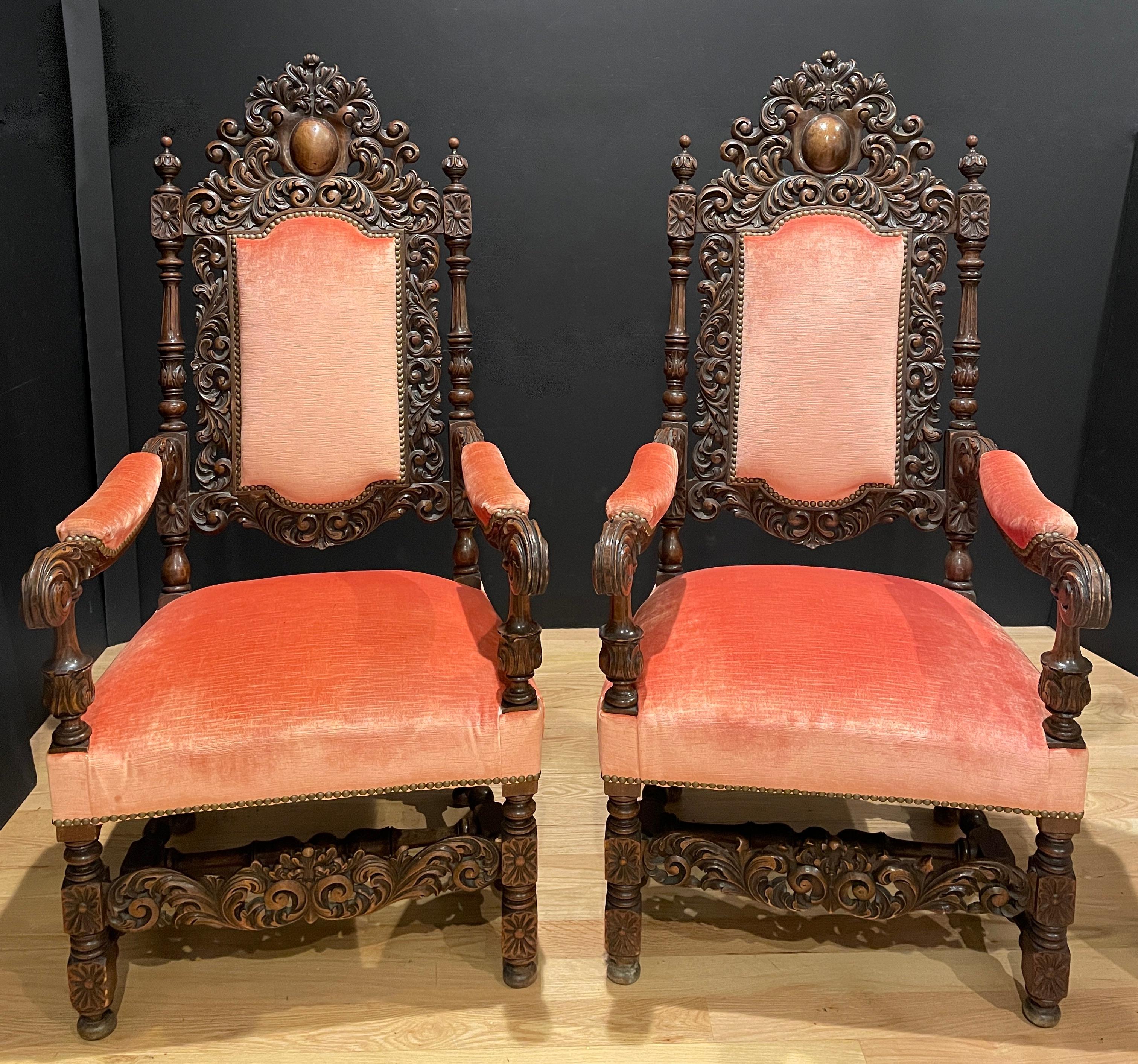 A fine quality pair of hand carved walnut Host and Hostess throne chairs. Upholstered in a coral tone velvet. Carving with a center cabochon form cartouche surrounded by floral scrolls and bottom carved apron.