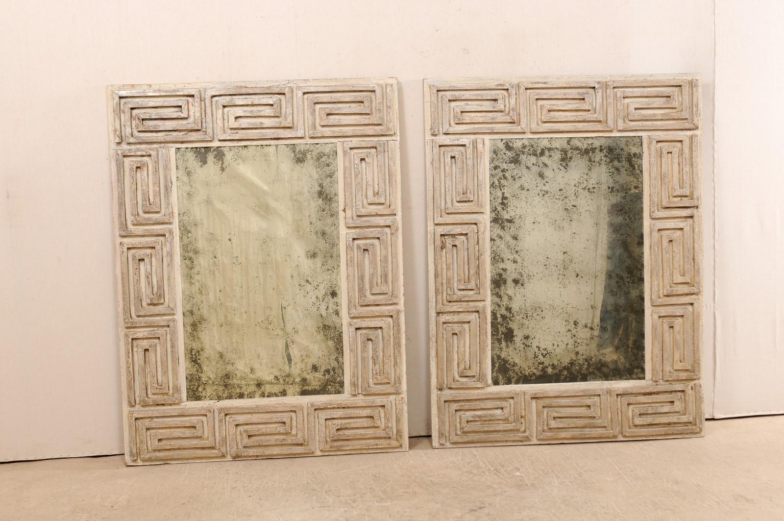 A pair of Greek key designed carved and painted wood mirrors from the 20th century. These antique mirrors, made in America, feature a thick, rectangular-shaped surround which is carved in Greek key design, with antiqued glass at each center. The