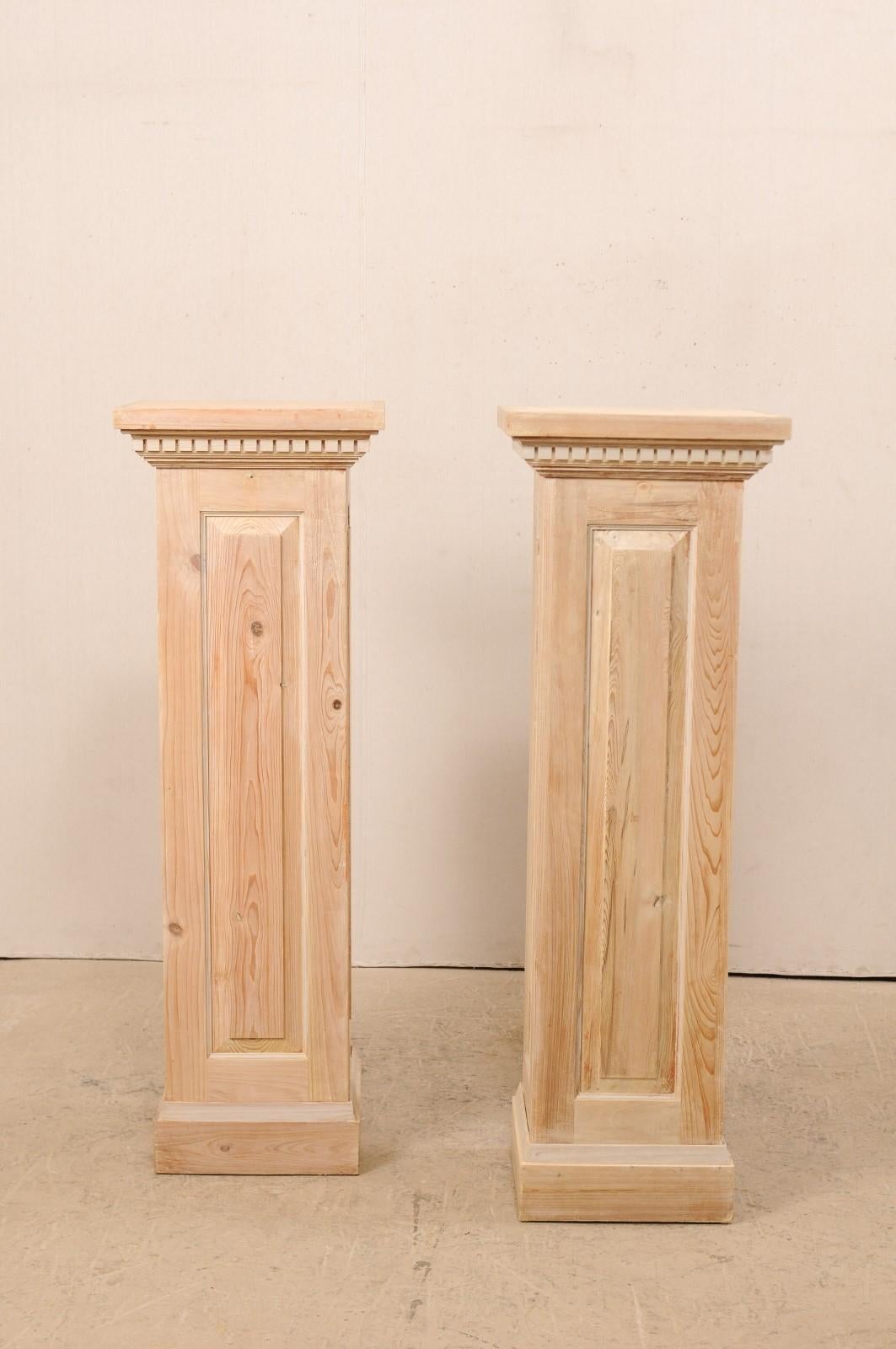A pair of vintage American carved wood pedestal columns. This pair of bleached wood columns feature squared bodies with a single raised panel at each side, stacked molding just beneath the overhanging square-shaped top, adorn with a row of dentil