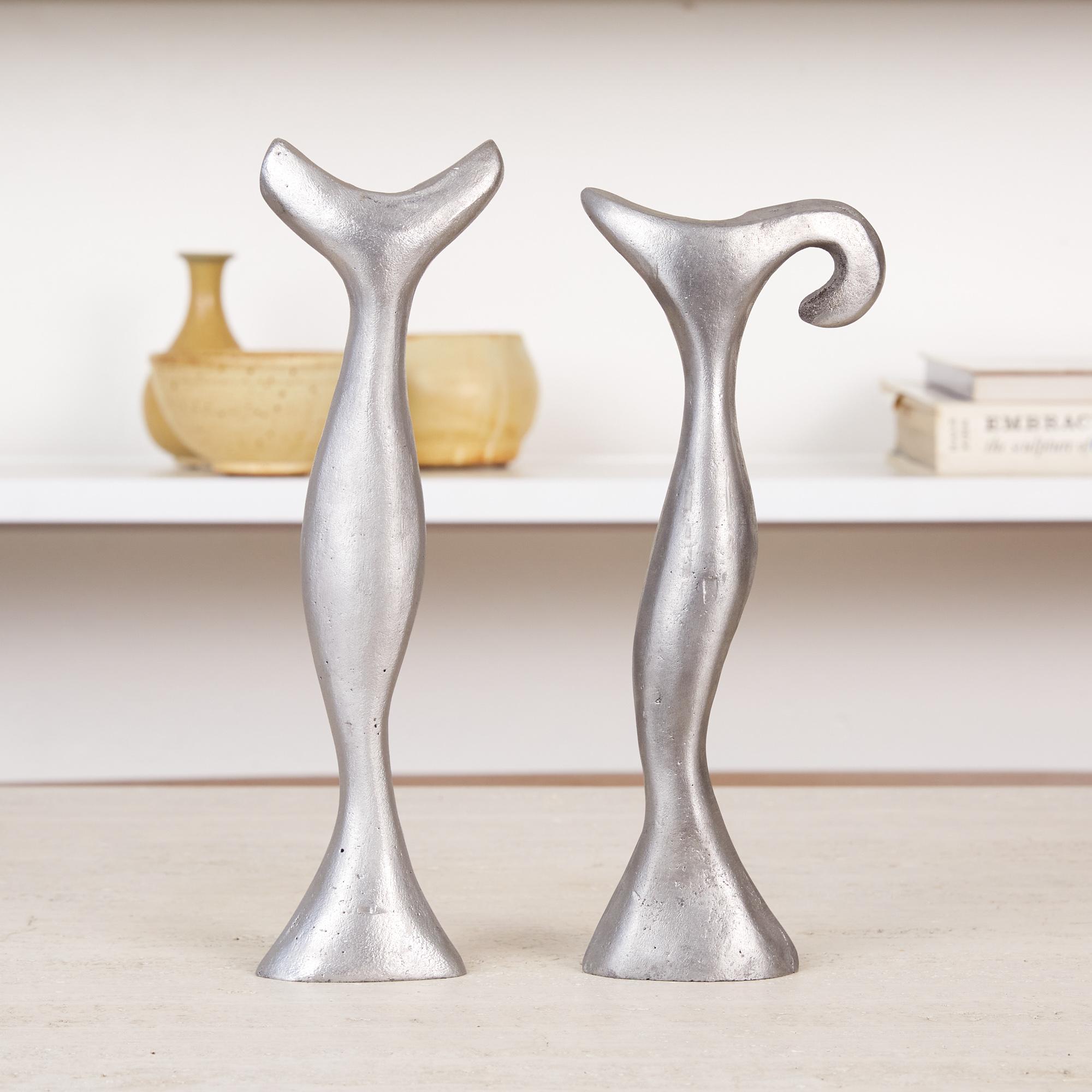 The pair of cast aluminum candlesticks feature an abstract form that goes up into a curvaceous opening that houses the candles. Small bubbles and slight imperfections left over from the casting process peep through the satin polish surface that