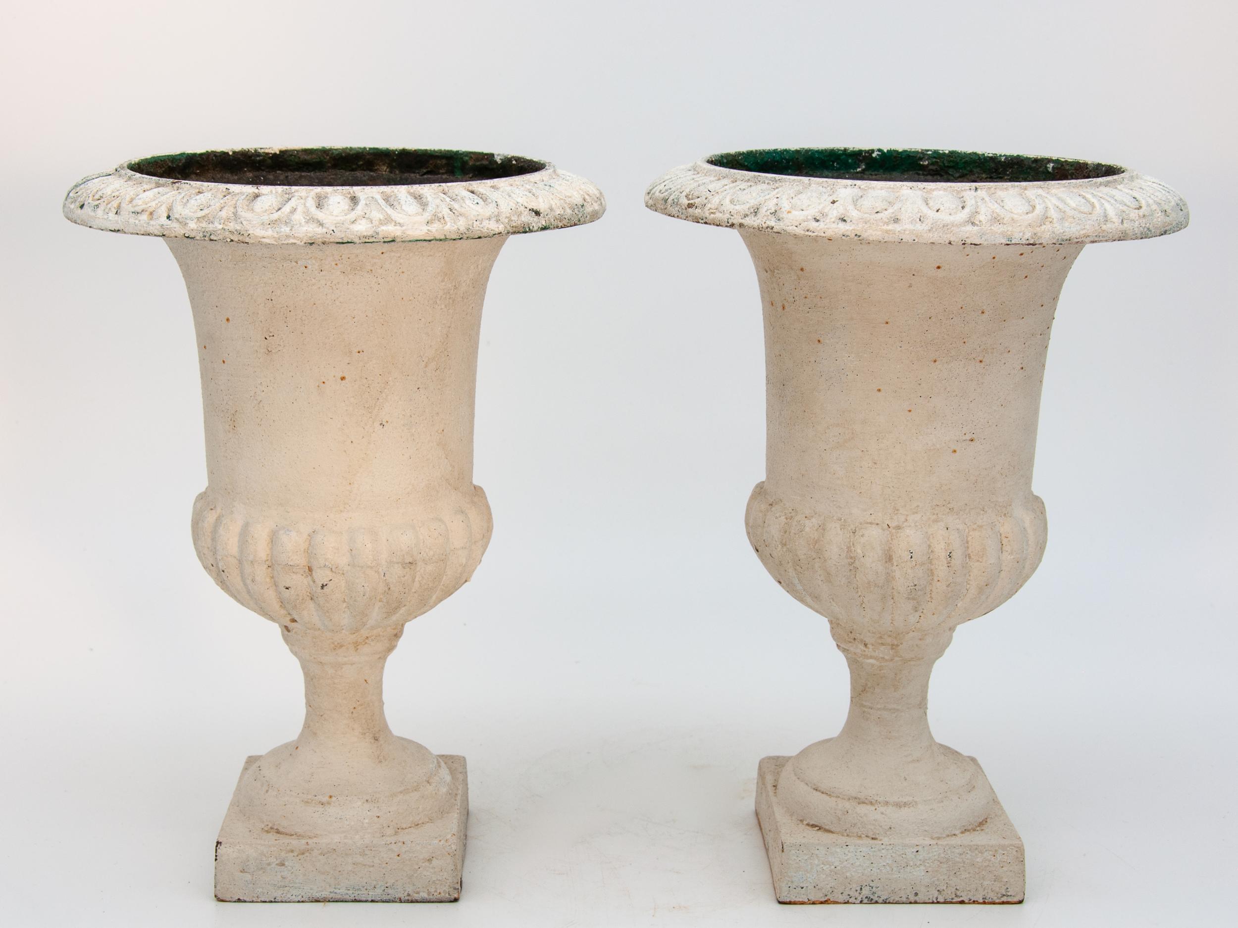 A pair of cast iron garden urns in a tabletop size. The lip of the urn has an egg and dart pattern the base of the urn is a classic fluted style on a square base. Later paint shows patina.