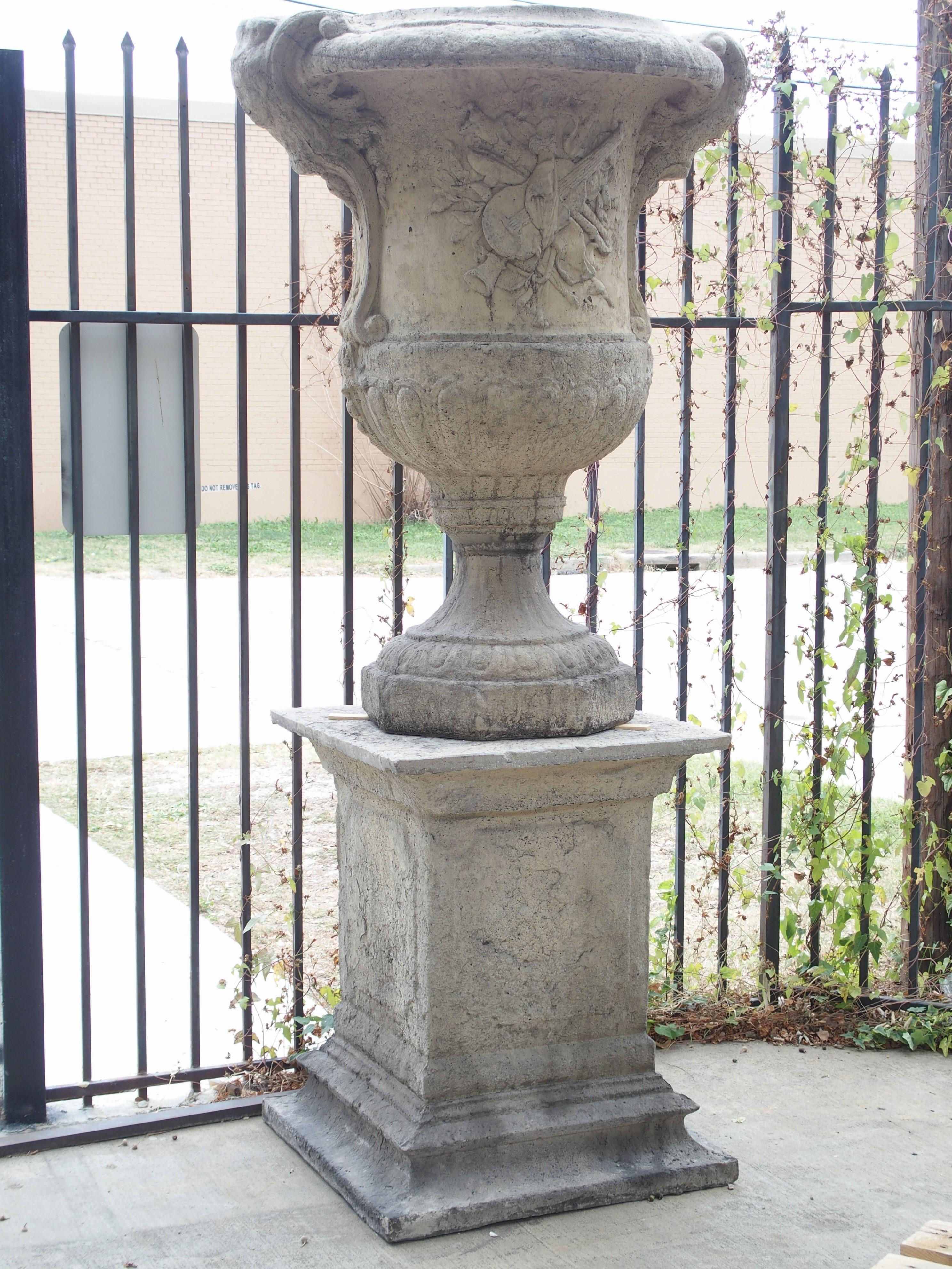 This pair of large cast stone vases on pedestals stands about 7 ½ feet tall and has a great worn texture and antique stone finish. Each vase is adorned with musical trophy decoration on both the front and back sides. The central element of the