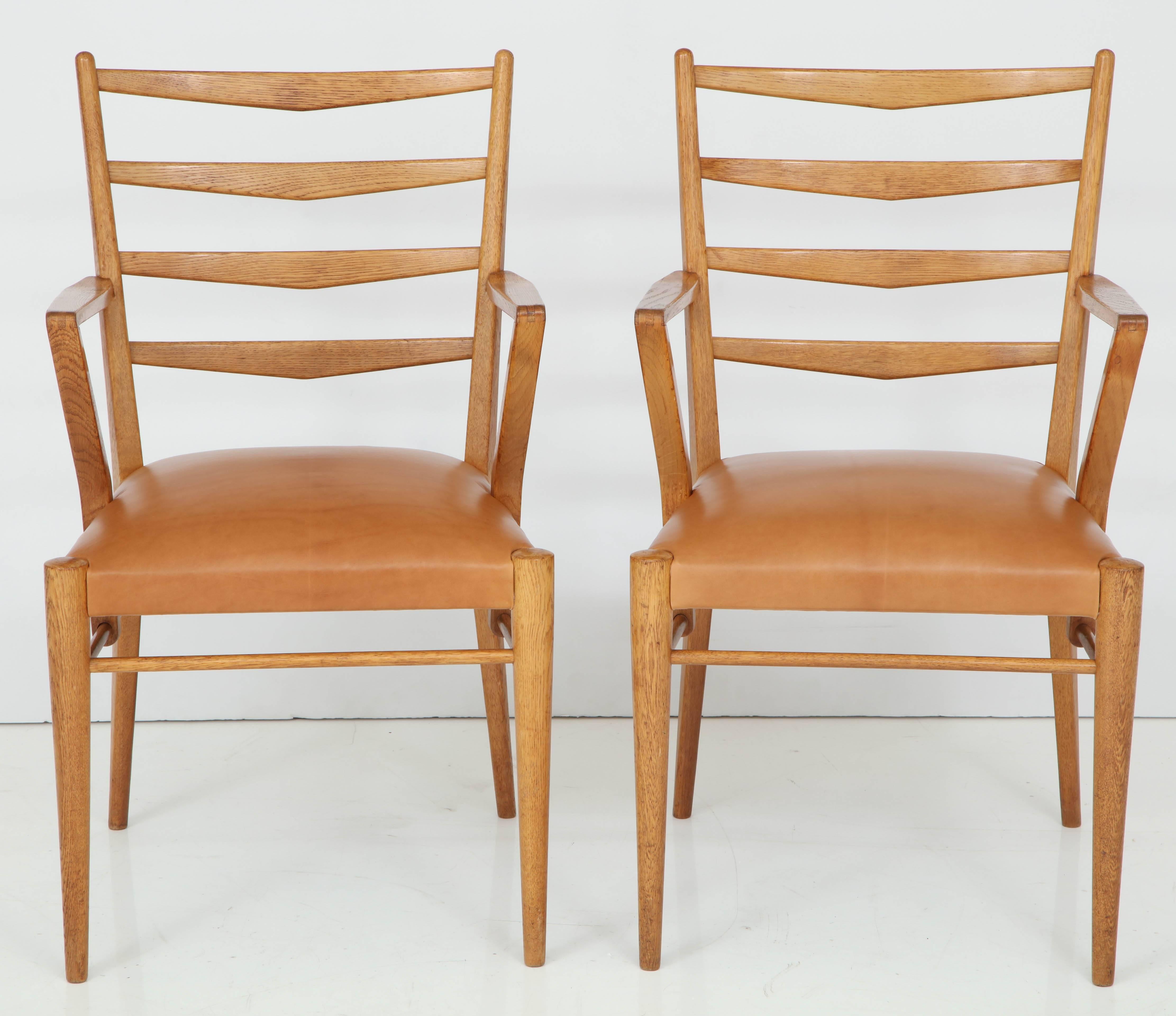 A pair of Dutch oak and leather open armchairs, circa 1950s, designed by Cees Braakman, each with ladder backs, triangular cutback armrests, an upholstered seat raised on circular turned and tapered legs.