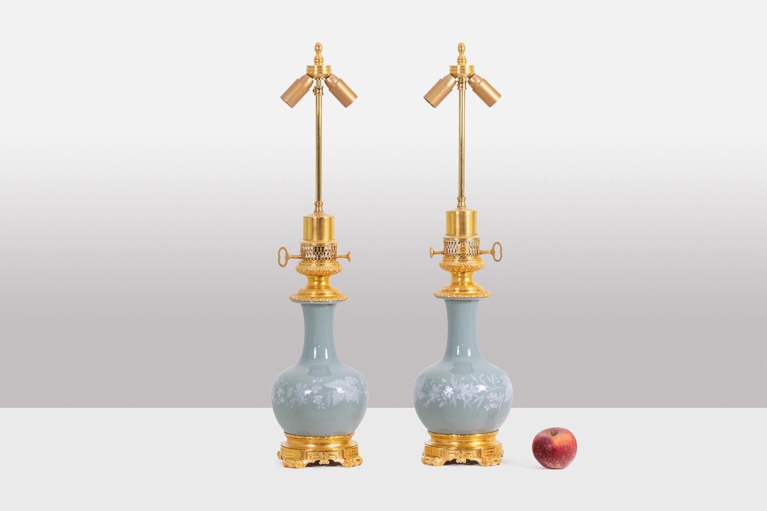 Paire de lampes de forme balustre en porcelaine de Céladon et bronze doré, la porcelaine décorée de fines fleurs blanches. Base circulaire et quadripode se terminant par des volutes, le haut du cadre décoré de godrons se terminant par une galerie