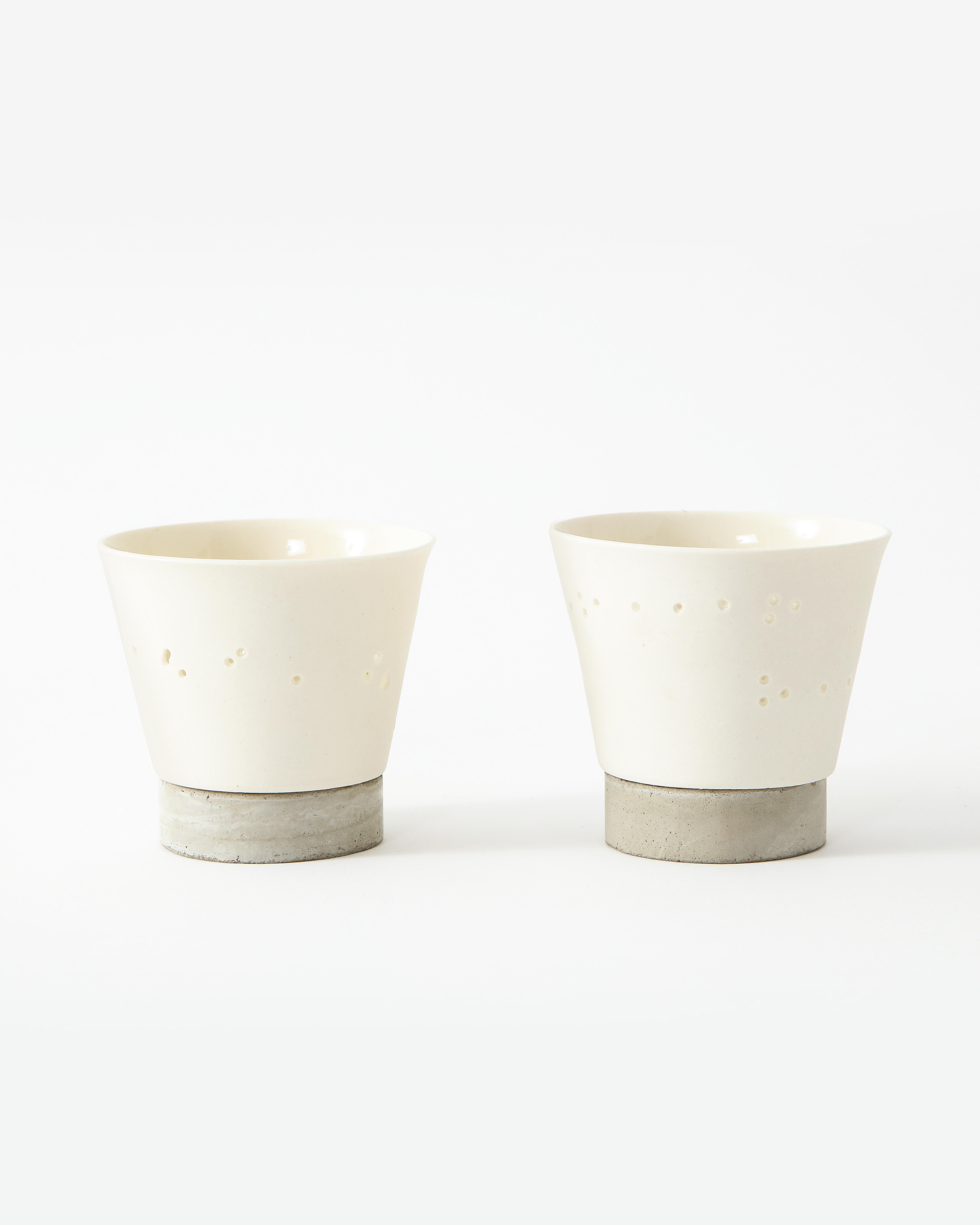 These soft white cups in thin ceramic are enhanced with small piercings that form a seemingly haphazard yet elegant design. Concrete bases finish off the bottom of the work.