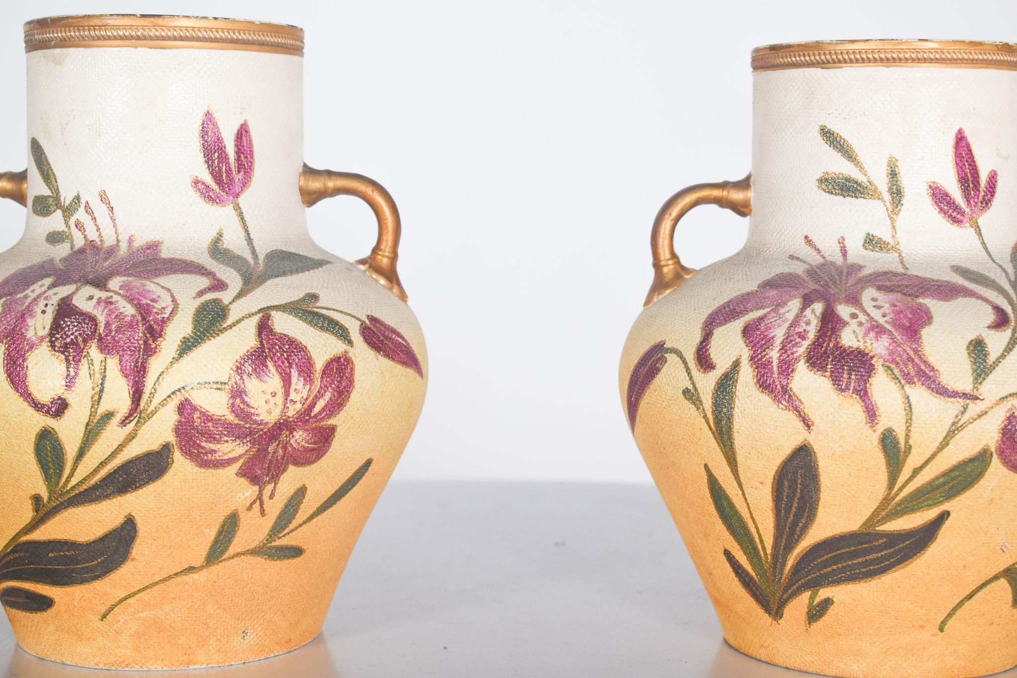 Paire de vases en céramique avec deux anses. Décor polychrome de motifs floraux, mettant en valeur des fleurs dans les tons pourpre et lie-de-vin, accompagnées de feuilles vertes. Les fleurs présentent des pétales allongés et texturés, ce qui donne