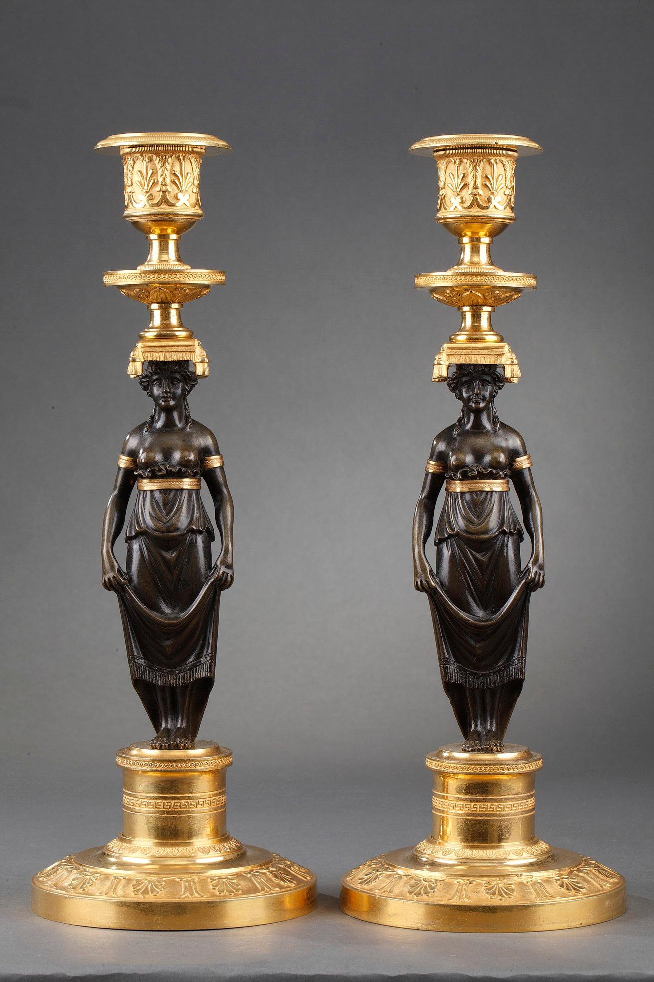 Beautiful Charles X pair of candlesticks in patinated and gilded bronze. The shafts present caryatids with bare chests whose drapery is retained at the waist by a gilded belt. They hold back their skirts to reveal their feet. They wear golden