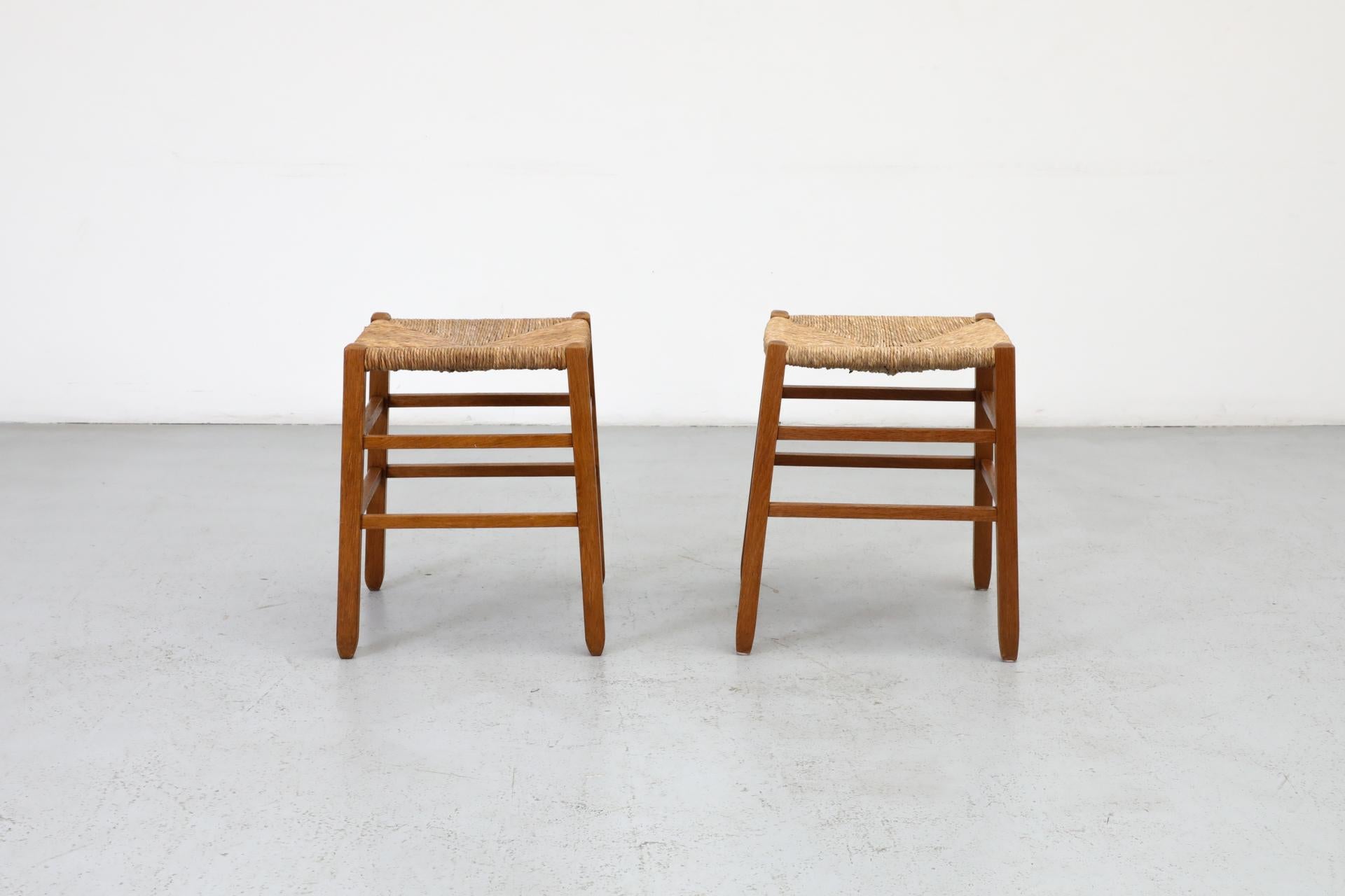 Pair of Charlotte Perriand inspired primitive oak stools with square legs and rush seats. In original condition with some patina and wear consistent with age and use. Other similar stools available and listed separately. Sold as a set.