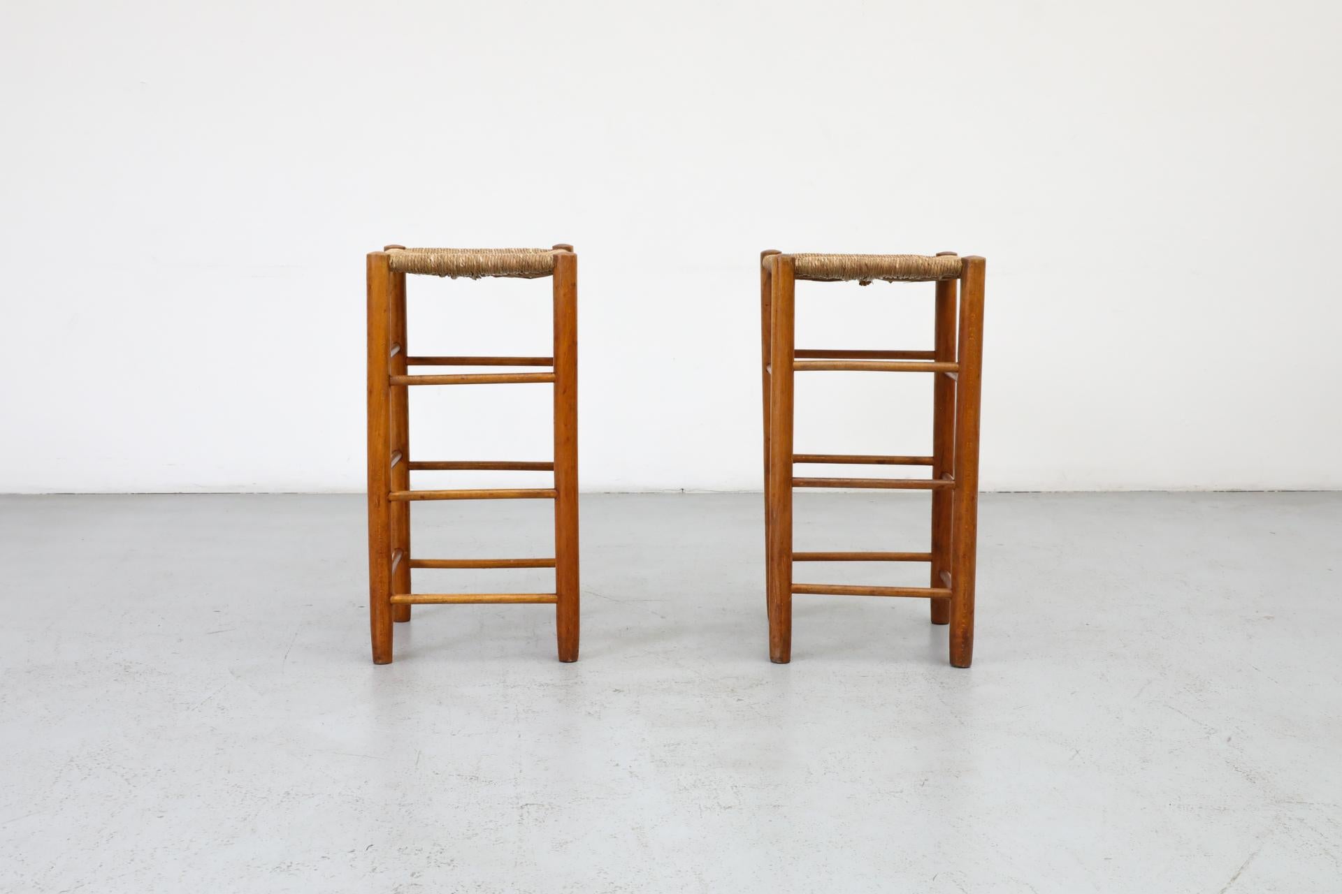 Set of two Charlotte Perriand style counter stools with woven rush seat and lightly stained pine frames. In original condition with visible wear and may have some rush breakage. Wear is consistent with age and use. Other similar stools available and