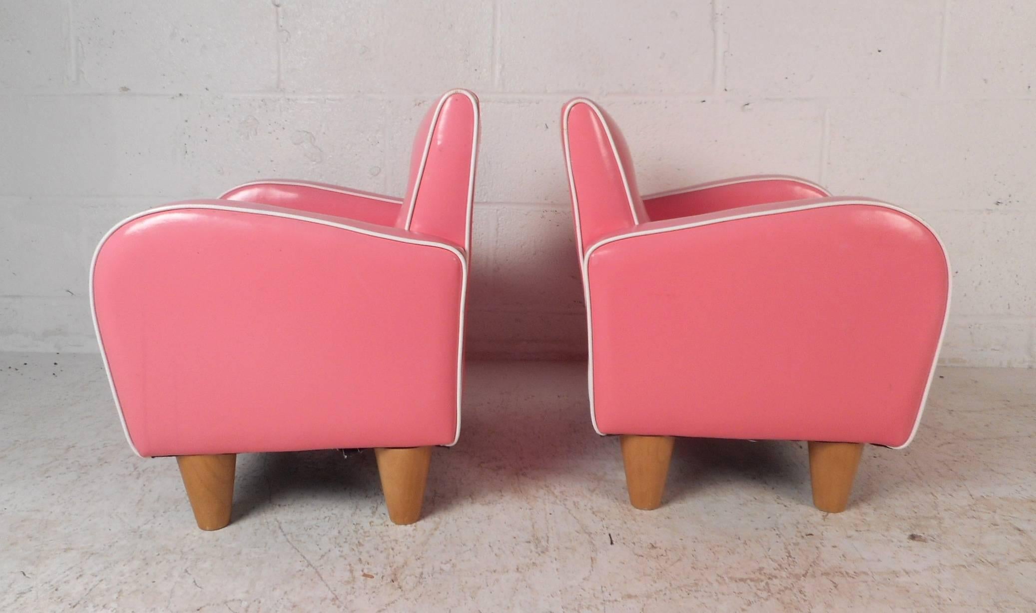 This wonderful pair of Art Deco style children's lounge chairs are covered in elaborate pink vinyl with high arm rests lined with white trim along the edges. These beautiful chairs have tufted back rests and sturdy tapered legs. This stunning pair