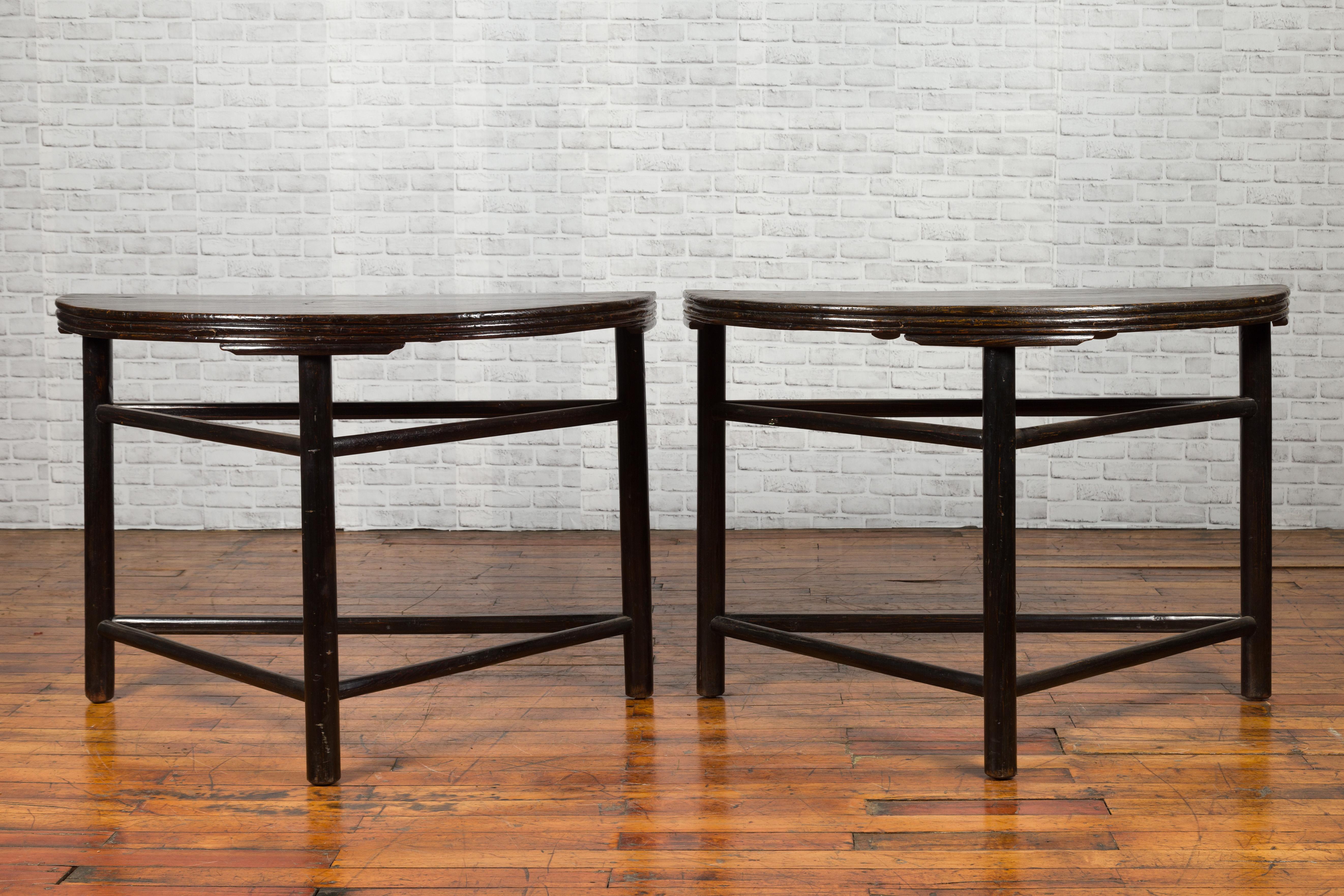 A pair of Chinese Qing dynasty period demilune console tables from the 19th century, with dark patina and side stretchers. Created in China during the Qing dynasty, each of this pair of demilunes features a semi-circular top sitting above three