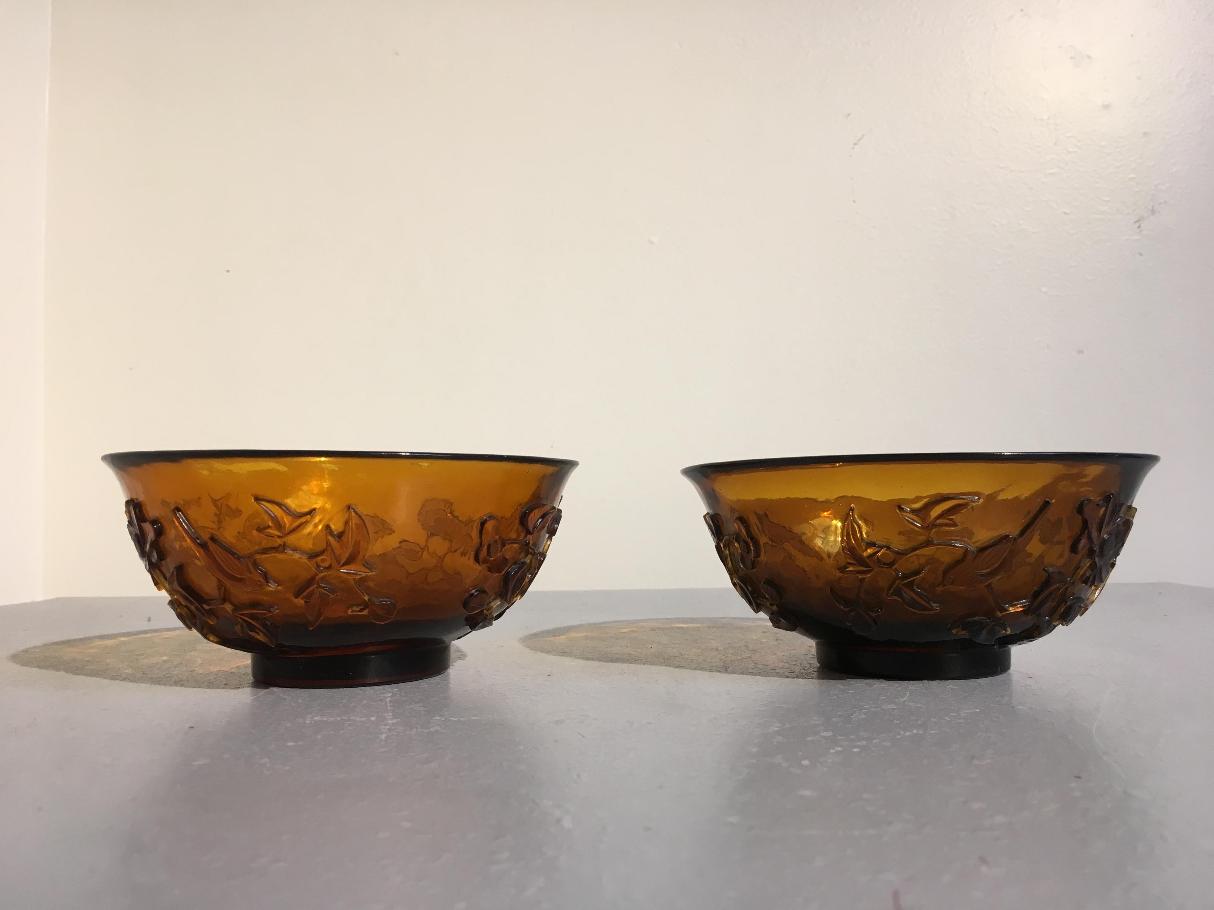A fine pair of late 19th century Chinese Qing dynasty carved Peking glass bowls in a lovely amber hue. 
The matching Peking glass bowls carved with opposing scenes of birds amongst flowering branches on a rocky outcrop. The amber glass bowls with