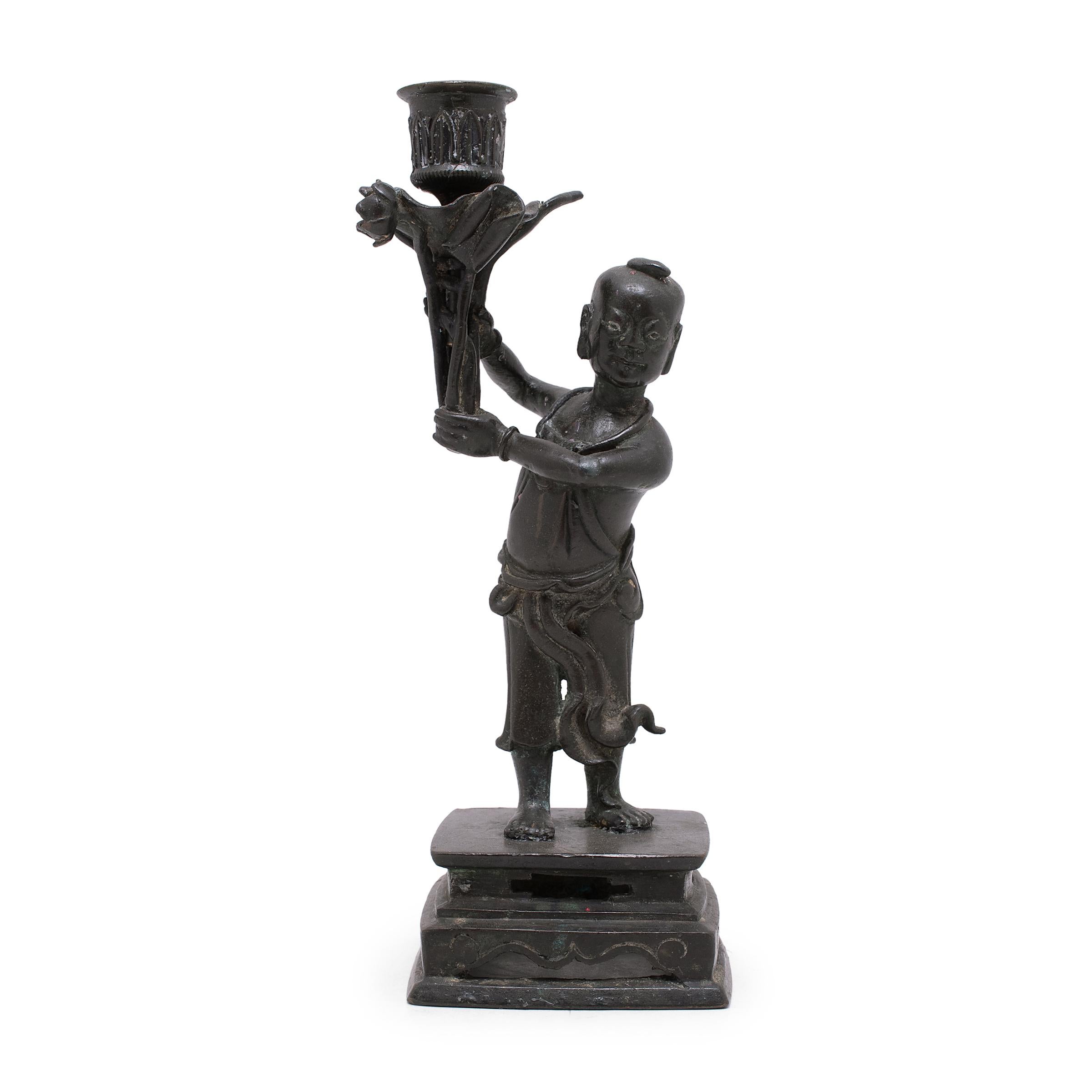 These early 20th century candlesticks are cast of bronze in the form of two young boys dressed in flowing garments and standing on square pedestal bases. Each boy holds a dish for taper candles overhead, tucked within a cluster of lotus leaves,