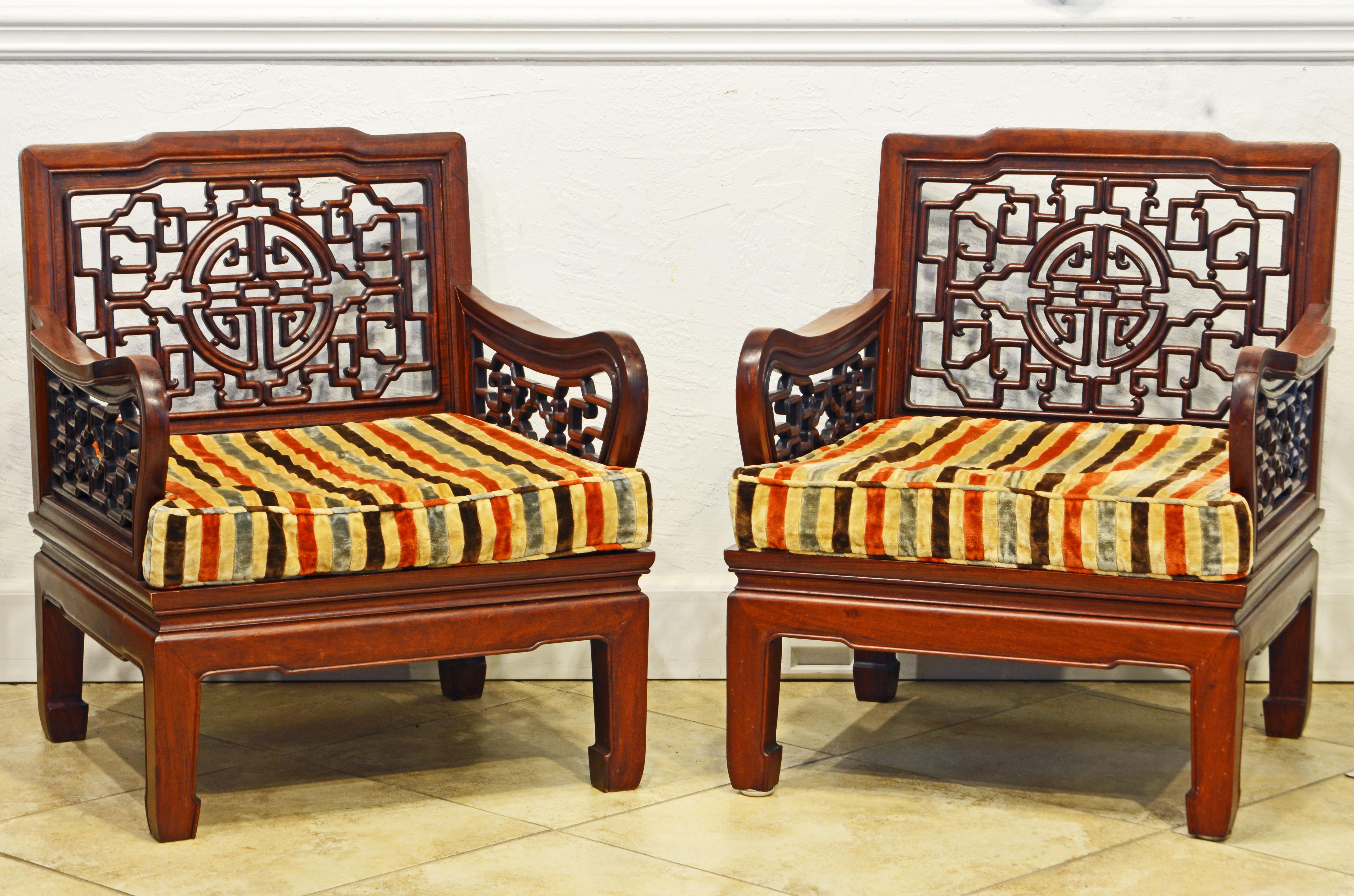 An inviting pair of colonial style carved mahogany club or lounge chairs with cushions dating to the mid-20th century and most likely made in Hong. Kong. The chairs which are reminiscent of Ming furniture feature backrests and arm rests with