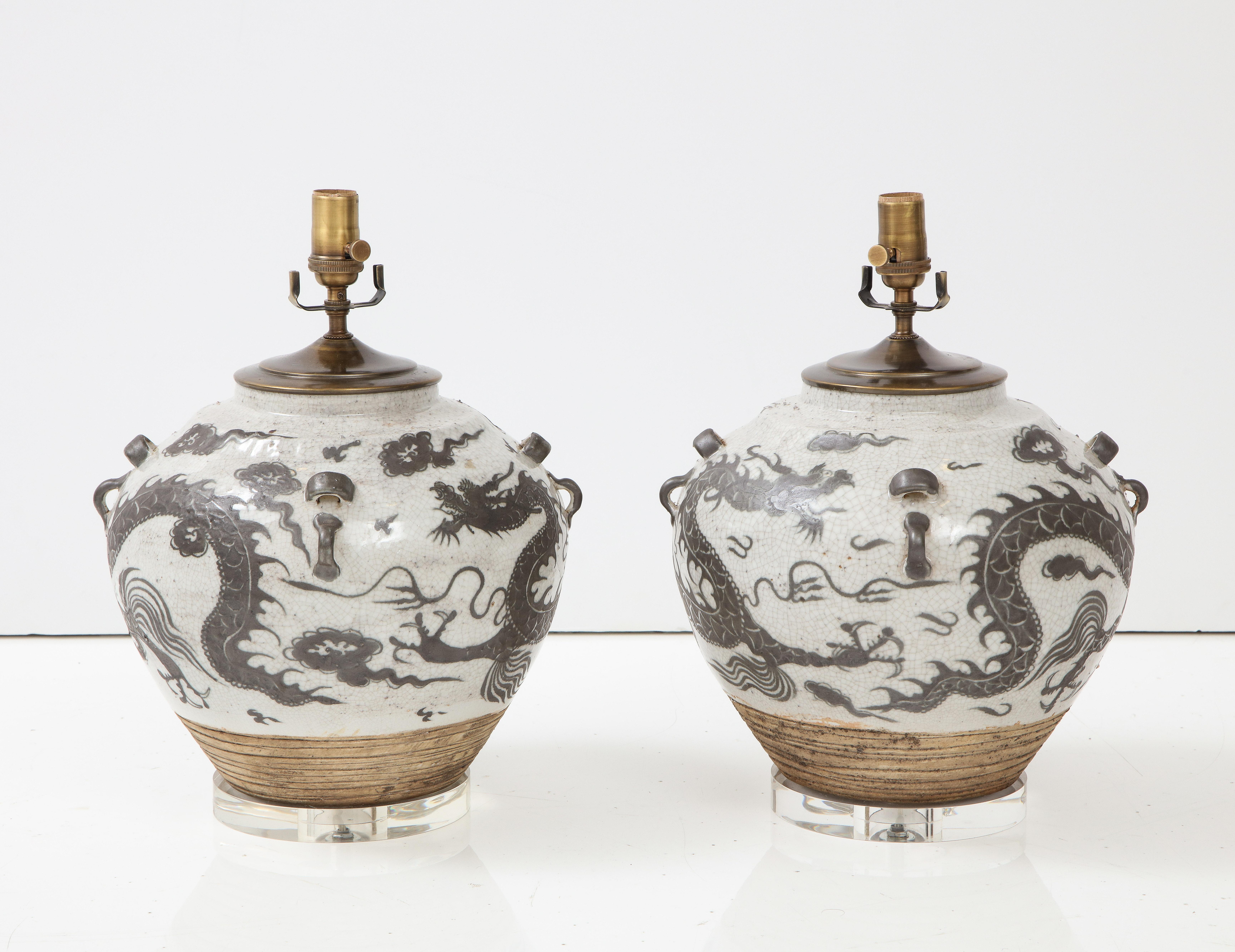 A pair of Chinese export jugs converted to lamps on a lucite base. The lamps have a unique shape that sets them apart from other lamps and they are decorated with a dragon motif in a deep gray color.