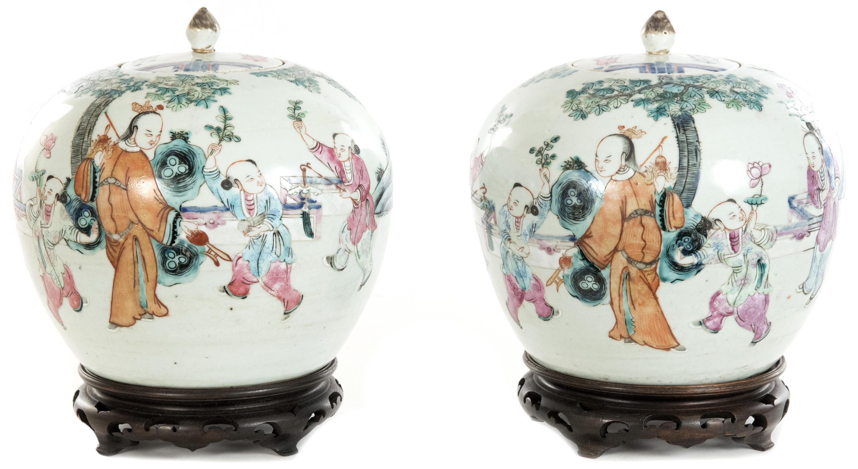 A pair of Chinese famille rose ovoid ginger jars, having a teardrop finial atop a circular lid, the white glazed body covered in round with depictions of children and an elder in a garden landscape, and mounted on a pierced wooden base. The