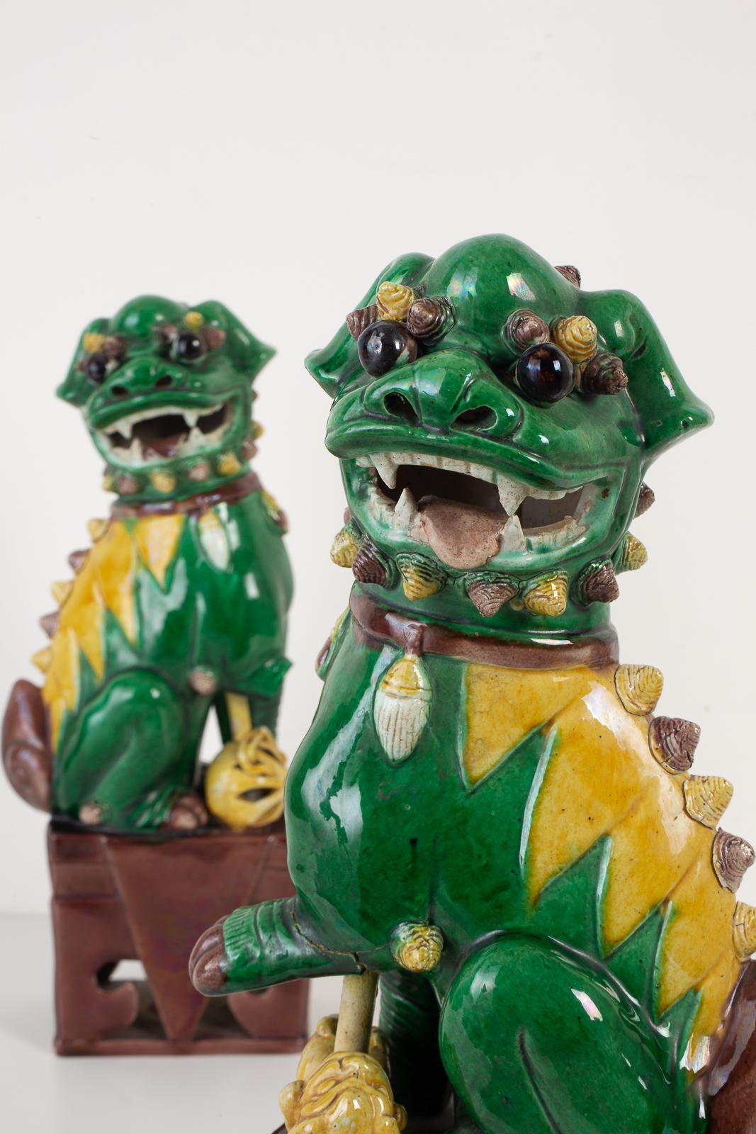 Fierce pair of ceramic Chinese Foo dogs on stands depicted sitting. Made in a beautiful famille verte green with bright yellow accents. Large size standing 14 inches high. One has a ball, and one has a cub under its arm. One has a damaged arm and