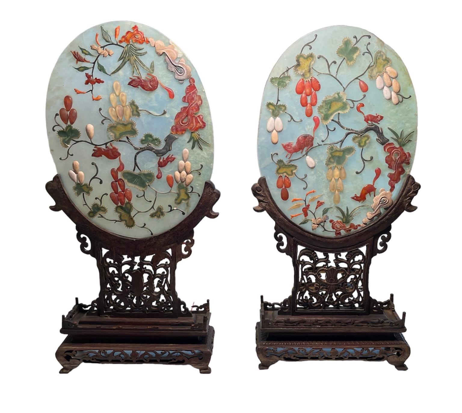This is a 19th century pair of Chinese hand carved oval jade and hard stone table screens with wood stands. They depict a semitransparent large egg shaped jade plaque adorned with a relief of grapes vines like branches of flowers made of agate, jade