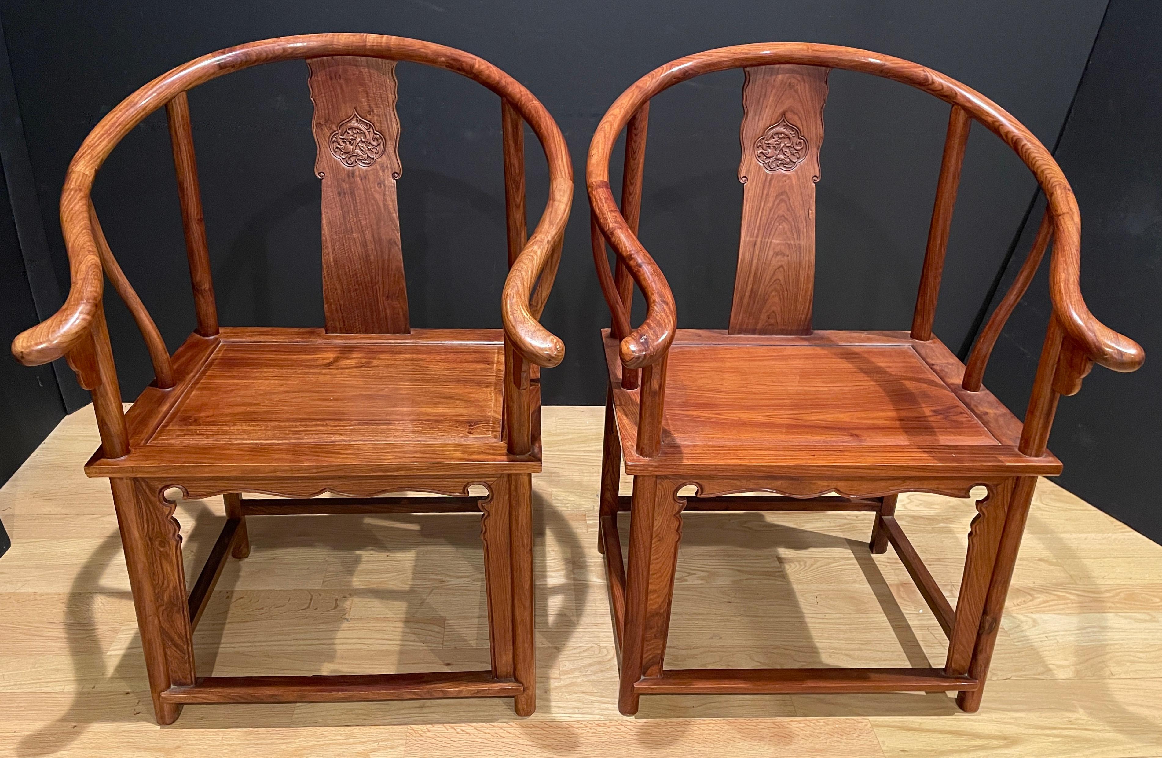 A pair of Chinese hardwood horseshoe back armchairs, each curved back with downswept arms and outswept hand grips, centering on an elegant shaped carved splat, flanked by cylindrical stiles continuing to undulating arm supports. The rectangular seat