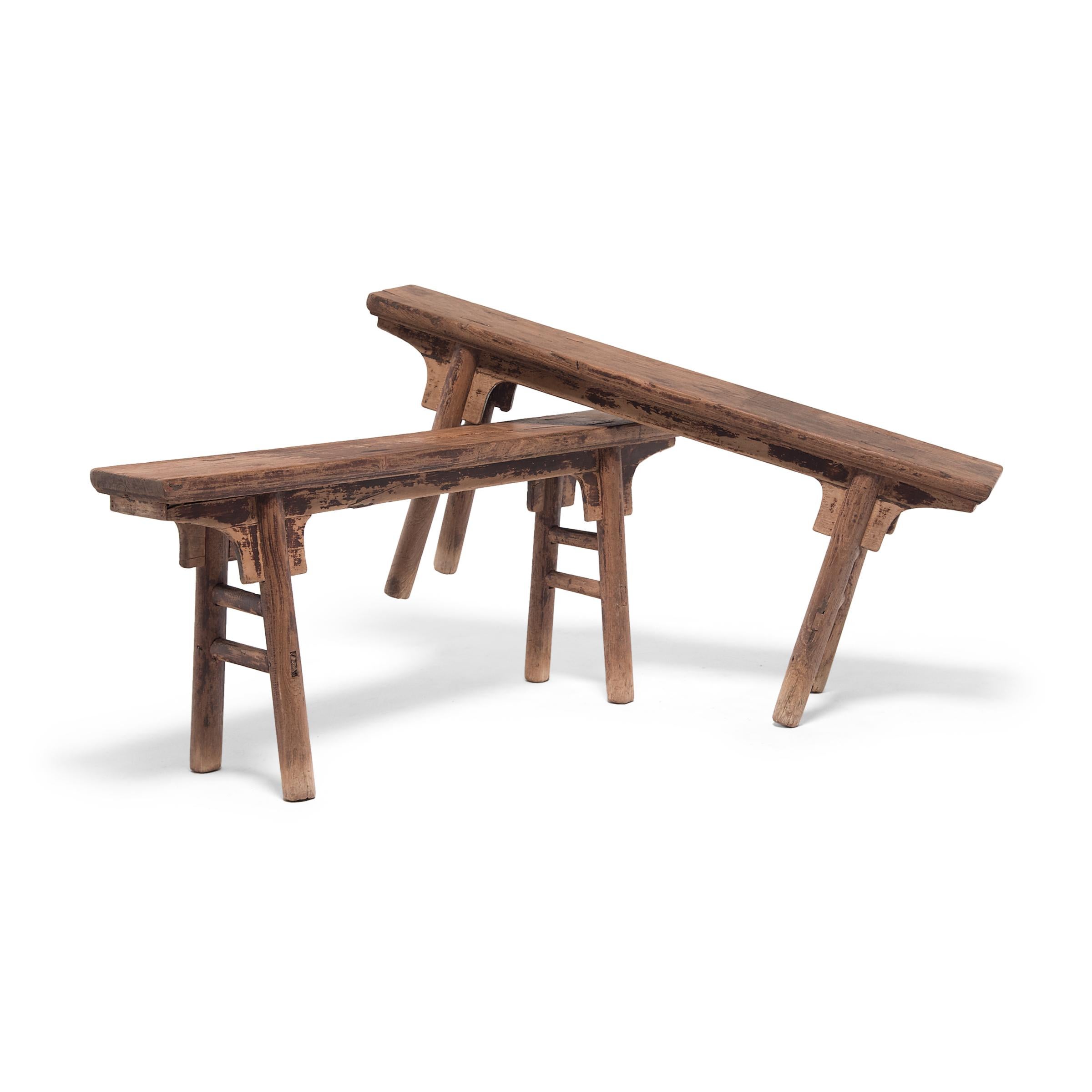 Dated to the early 20th century, these provincial benches from China's Hebei province feature a plank-top two-person seat supported by splayed round legs with double stretcher bars. The original dark finish has faded over time for a weathered look