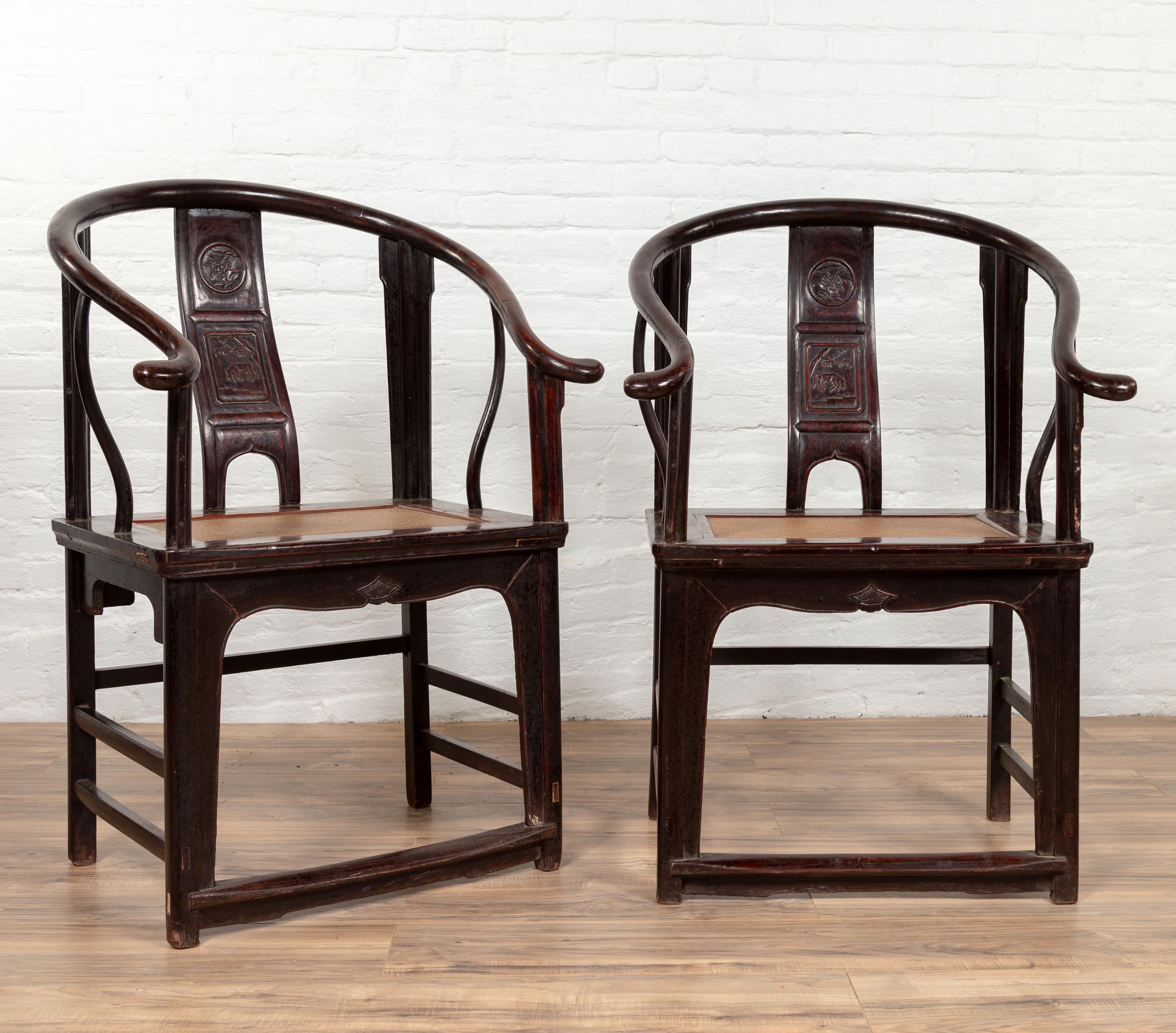 A pair of Chinese antique Ming dynasty style black wood horseshoe armchairs from the early 20th century, with carved backs and rattan seats. Each of this pair of black wooden armchairs features an exquisite horseshoe back, adorned with a sinuous