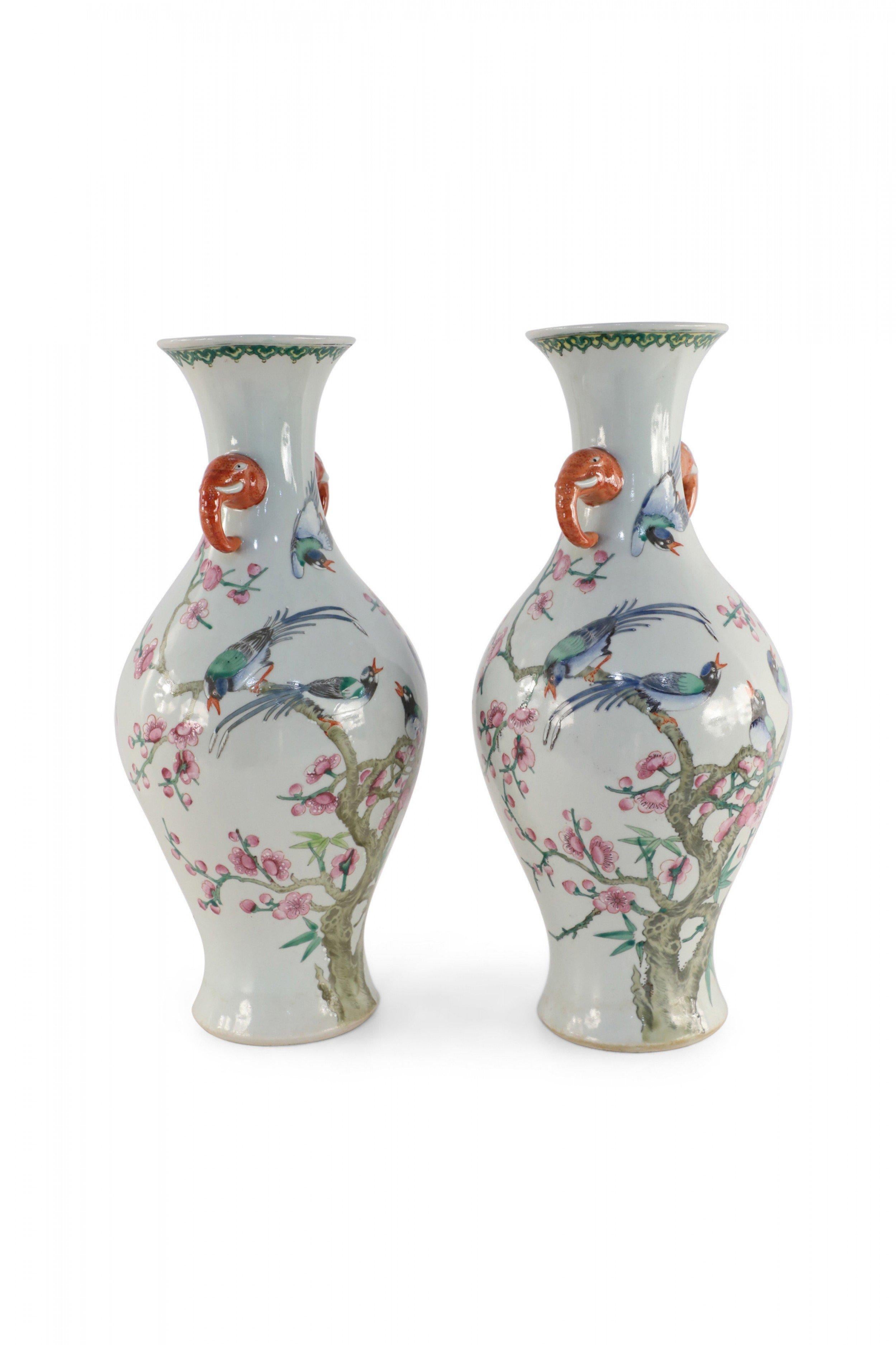 Pair of Chinese off-white urn-shaped porcelain vases depicting groups of birds perched on flowering branches of cherry blossom trees and one bird flying above, positioned between two orange, elephant handles that adorn the necks (priced as pair).
  