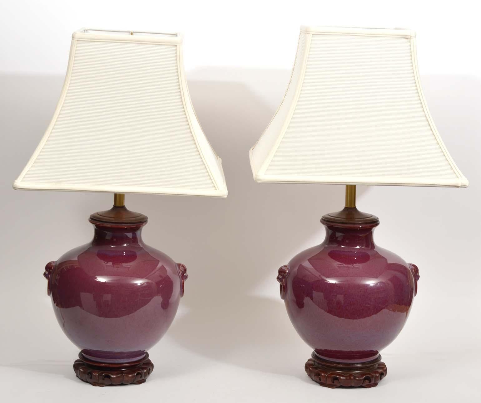 This pair of urn shaped Chinese oxblood glazed ceramic table lamps feature deep color surfaces accented by stylized lion head handles in haut relief. The rest on carved hardwood bases matching the circular wood lids through which the lamp pictures
