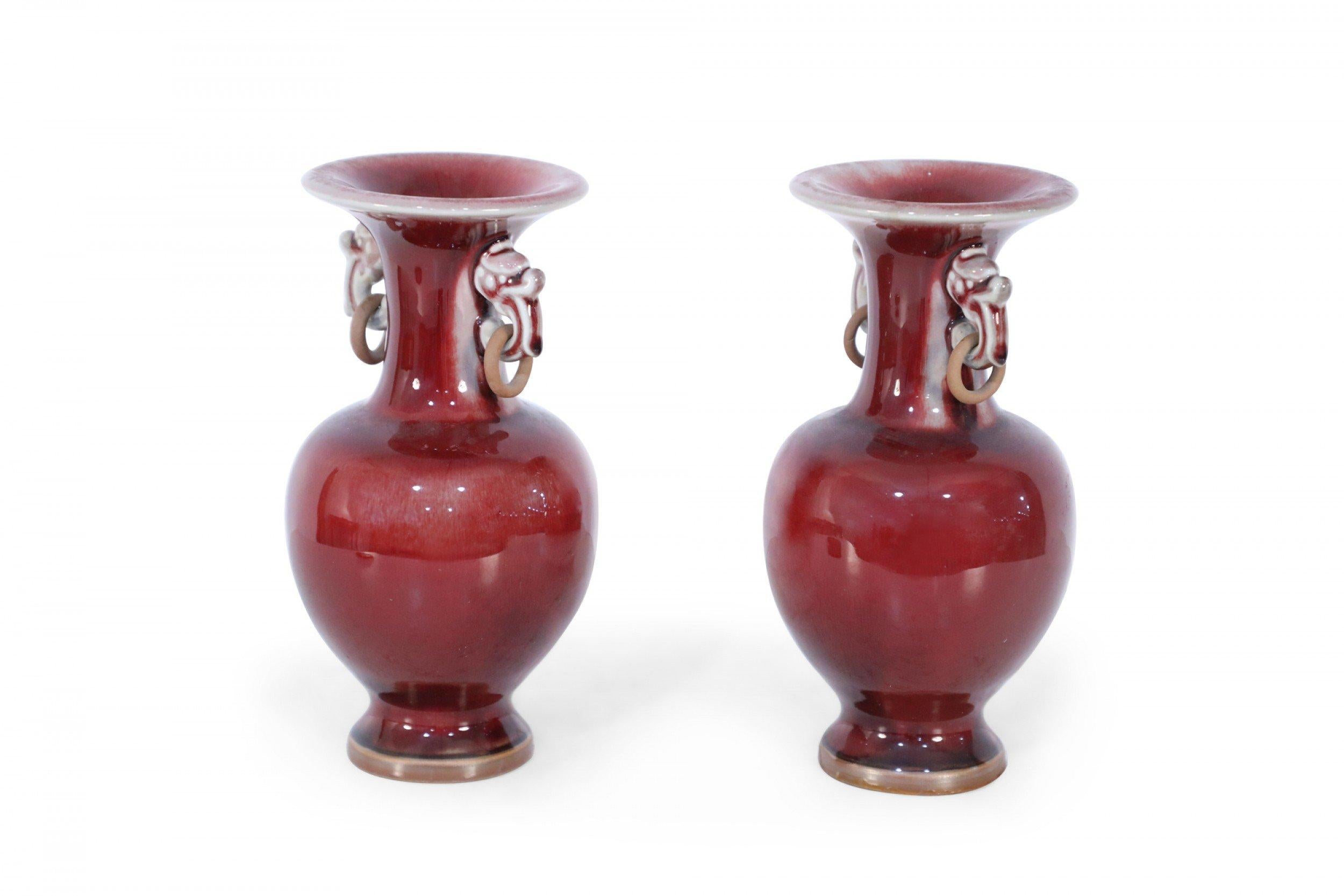 Pair of Chinese oxblood red vases with fluted necks, accentuated by figurative ears holding wooden rings, leading to bulbous lower halves, atop a small, round foot (priced as pair).
      