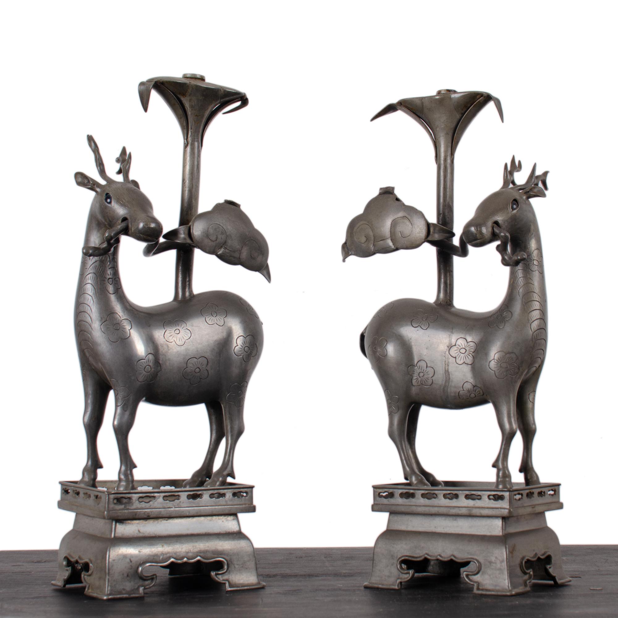A pair of Chinese figural pewter candlesticks in the form of deer, Qing Dynasty, circa 1880.  

Raised on rectangular plinth; the deer with inset cobalt glass eyes and decorated with incised flowers and holding lingzhi (reishi) mushrooms in their