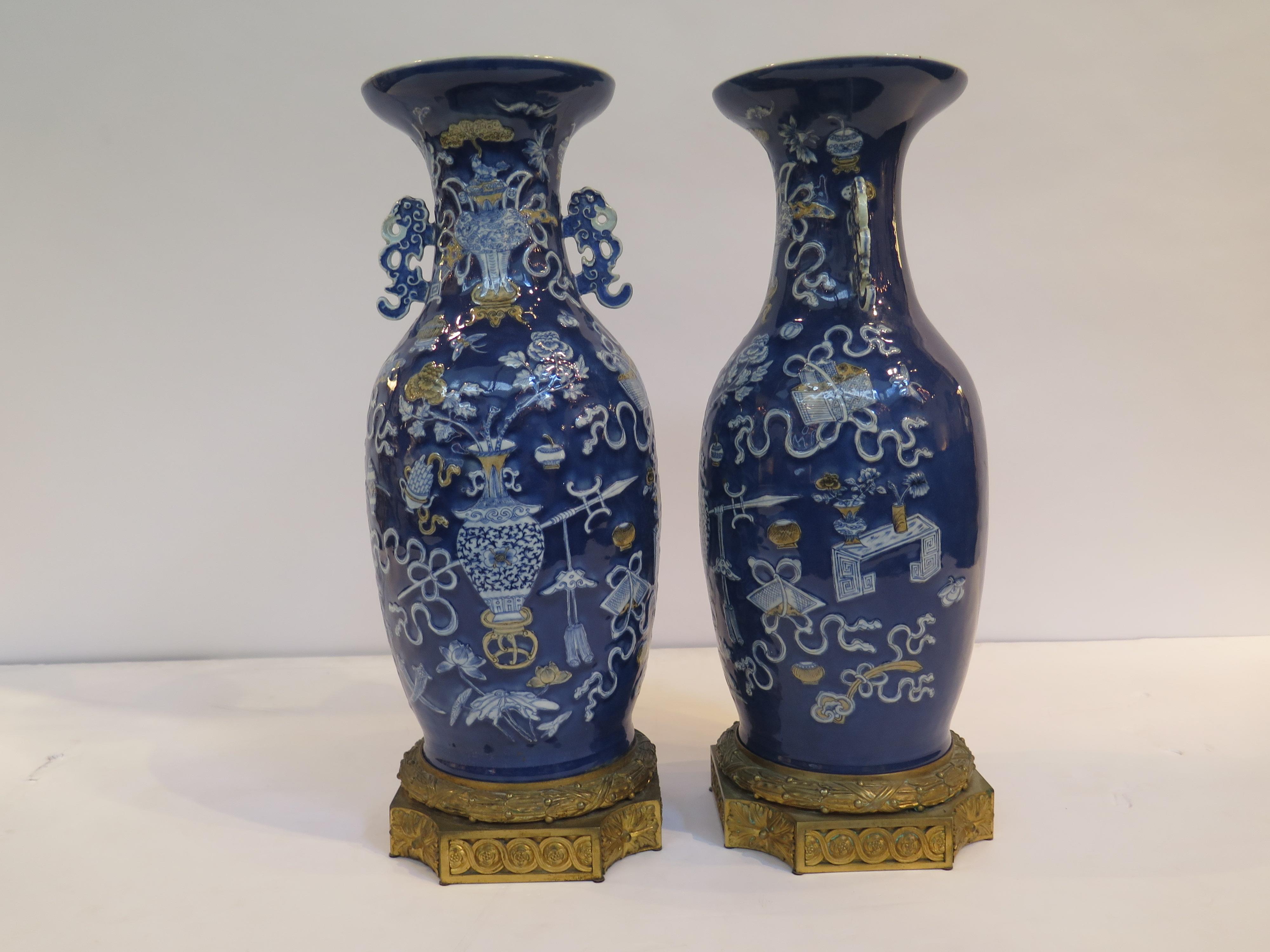A beautiful pair of Chinese porcelain vases mounted on elaborate French gilt bronze bases. The vases with a blue background have raised white floral bouquets in center and various treasures around them. Both with elaborate handles at neck. 

Handles