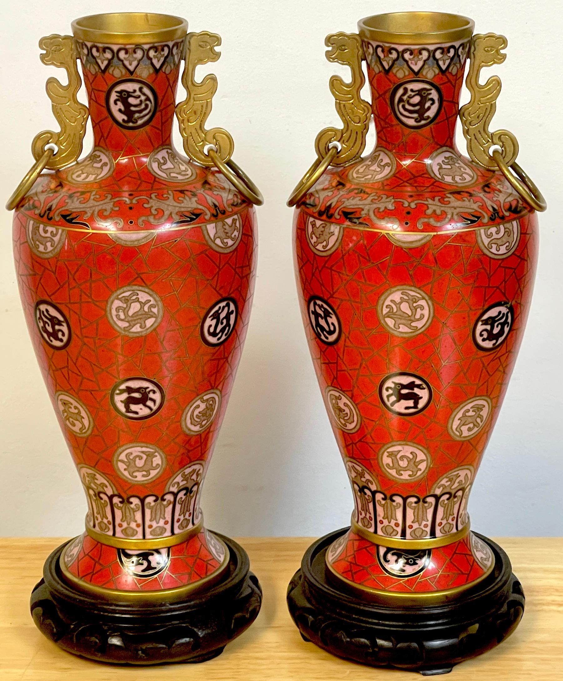 Pair of Chinese red cloisonné zodiac vases, with stands, each one with gilt brass phoenix bird ring handles, with 'cracked ice' and floating animal medallions. Complete with associated pair of carved hardwood stands. 
Unsigned 
Vase alone measures