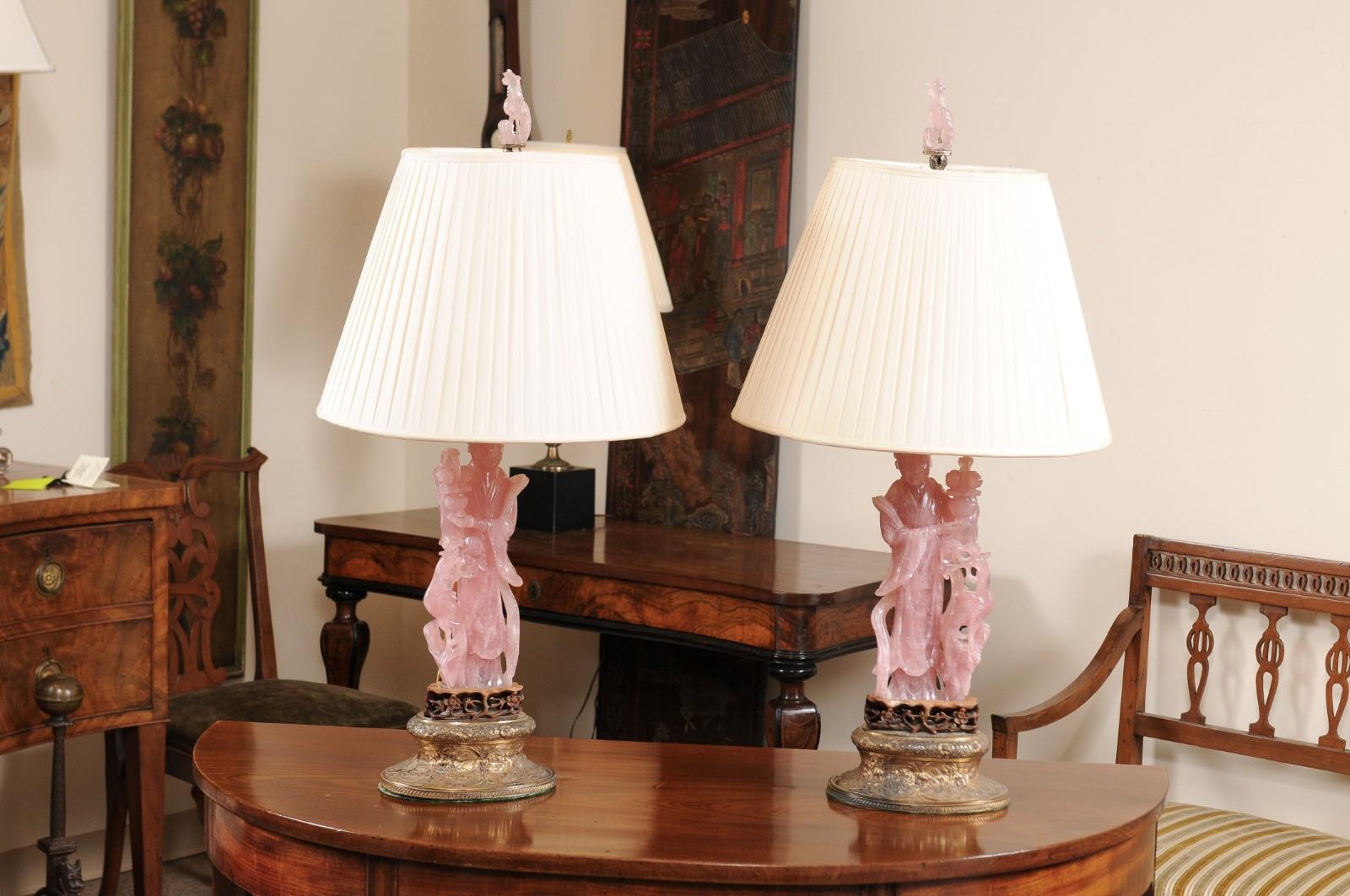 The pair of Chinese rose quartz lamps of female figures holding floral vases and phoenixes, mounted carved wooden stands with oval gilt metal engraved bases. The lamps with silk shades, rose quartz finial and recently wired for USA electricity.
